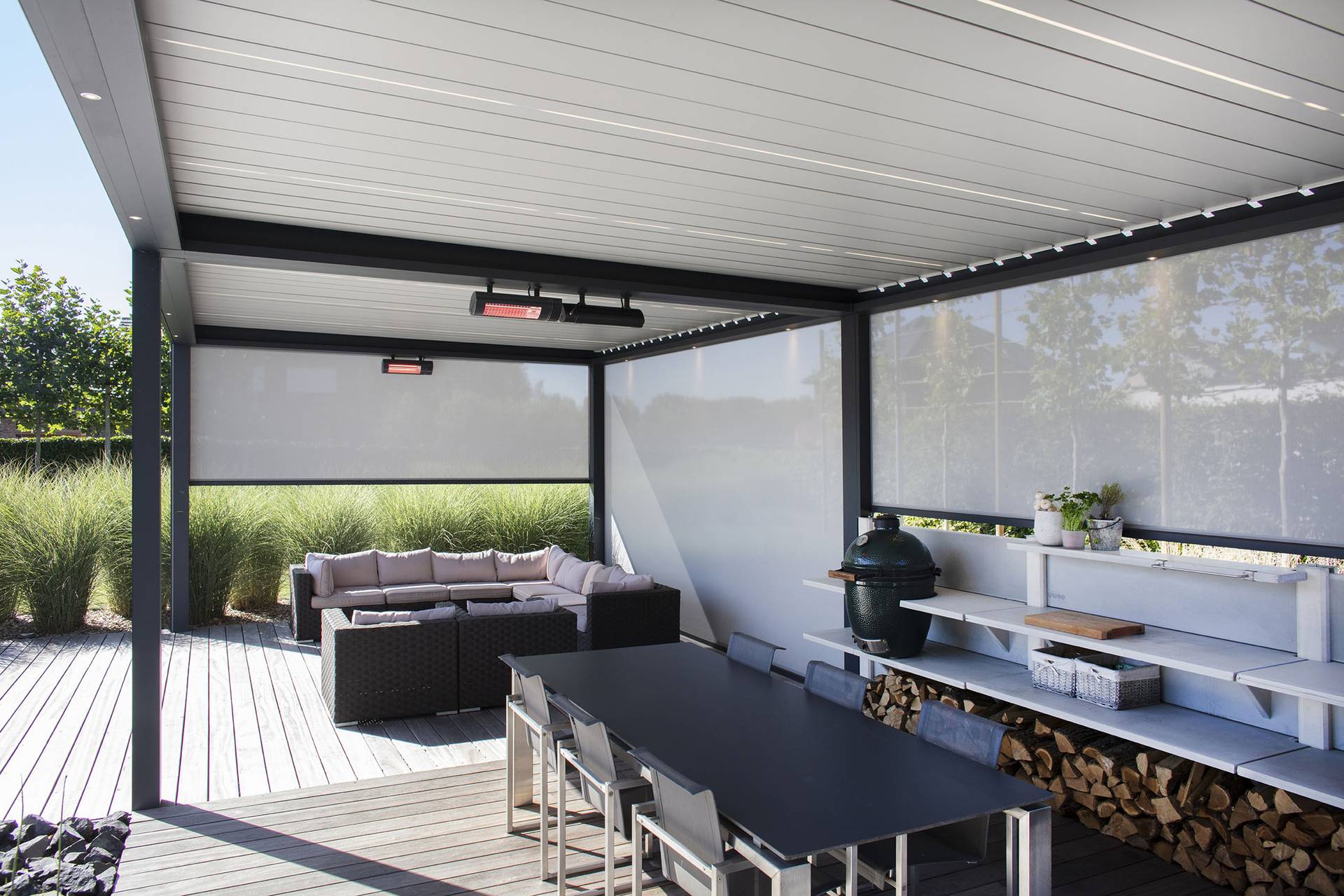 Aluminium pergola with tilting louvred roof, integrated heaters and lowered ZIP‑screens, shading an outdoor dining table and lounge area.