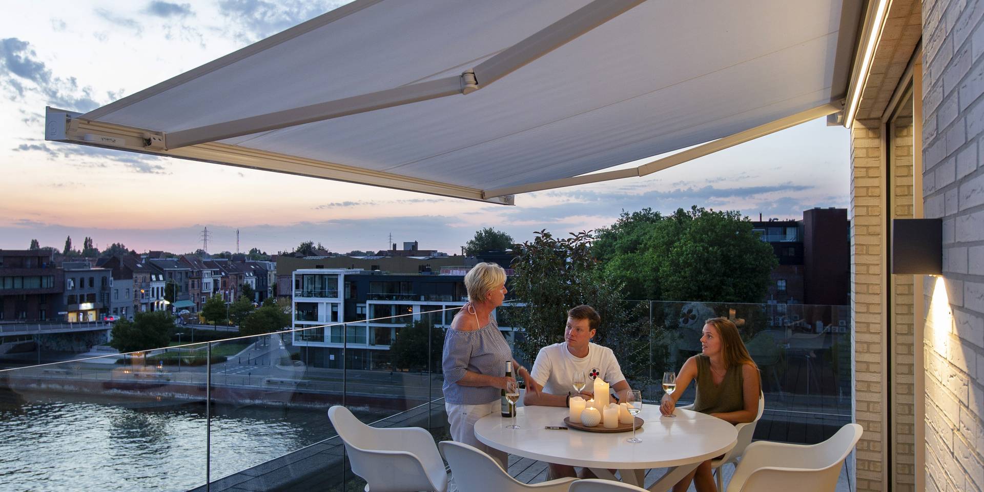 Aluminium awning extending over a balcony dining area with glass balustrade, overlooking the waterfront at sunset.