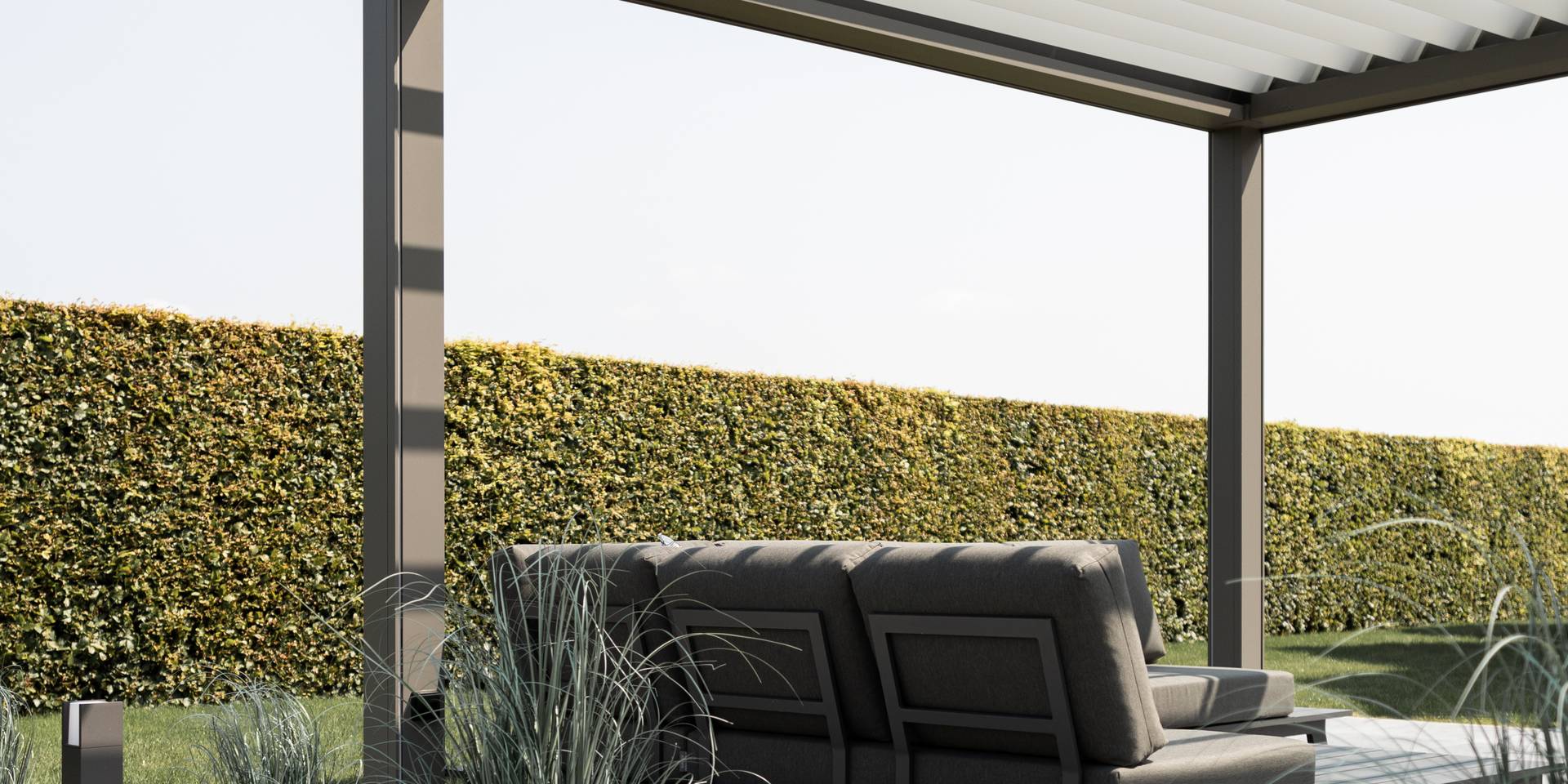 Espace lounge extérieur sous une pergola en aluminium à lames orientables, donnant sur une haie de jardin soigneusement taillée.