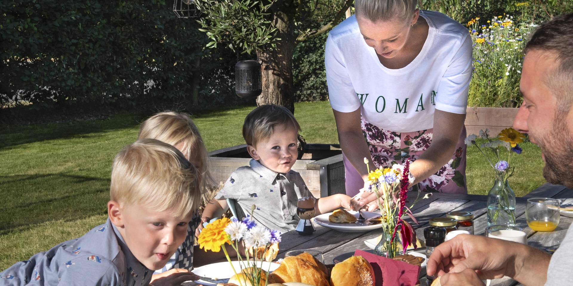 Scène de repas en plein air avec une table en bois garnie de nourriture et de fleurs dans un jardin ensoleillé.