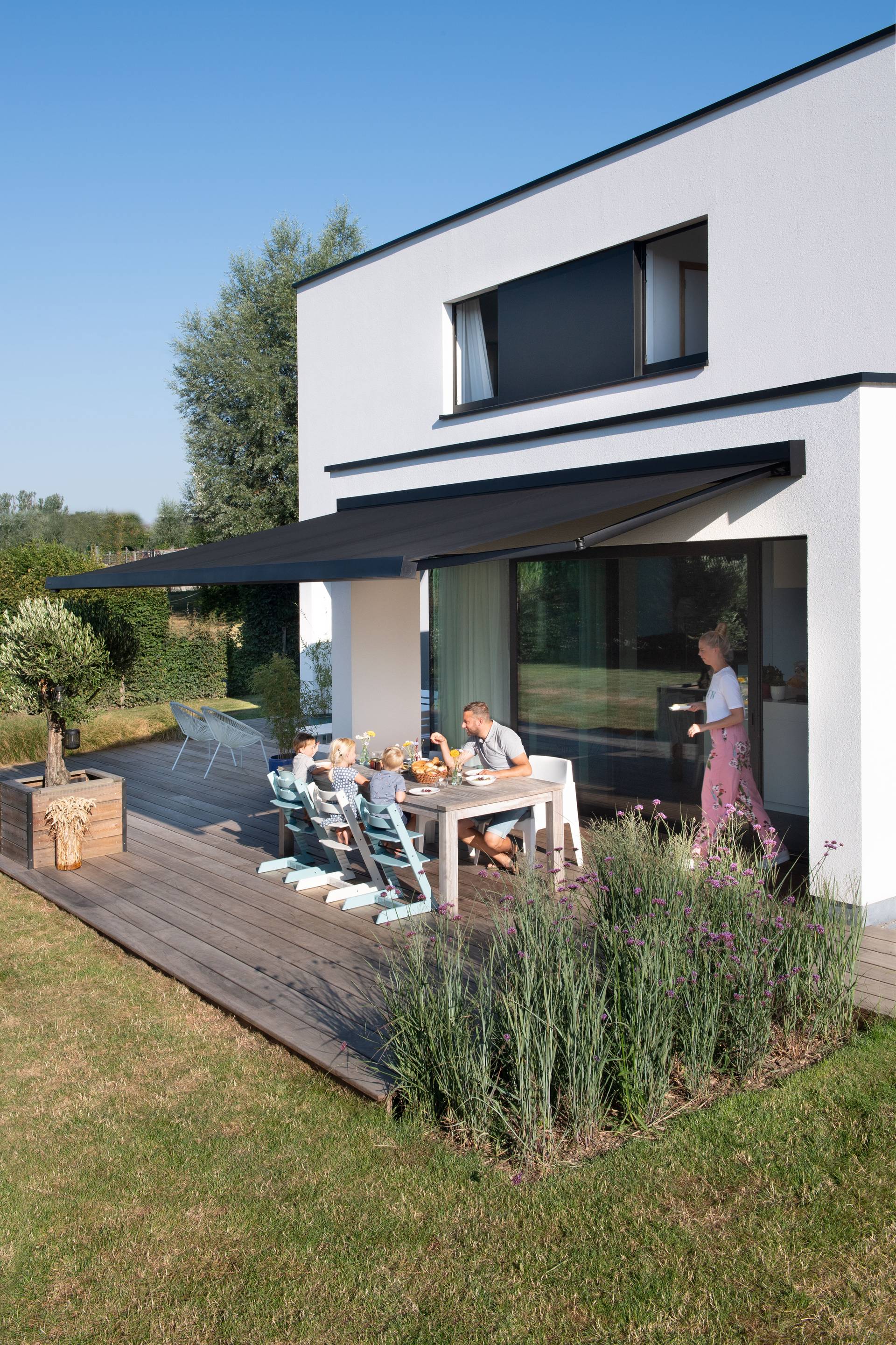 Maison moderne équipée d’un store banne rétractable en aluminium ombrageant une terrasse en bois avec espace repas extérieur, donnant sur un jardin paysager.