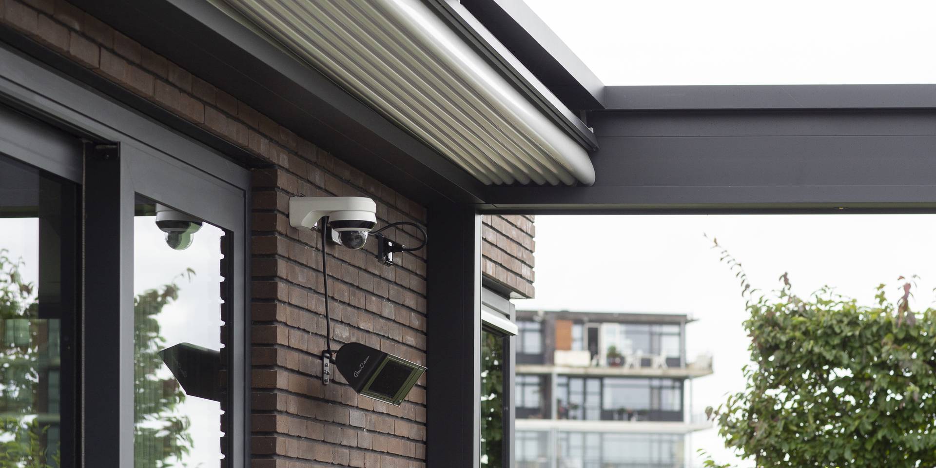 Gros plan d’une pergola avec toit en toile rétractable fixée à une façade en briques, montrant la cassette, les rails de guidage et un éclairage extérieur.