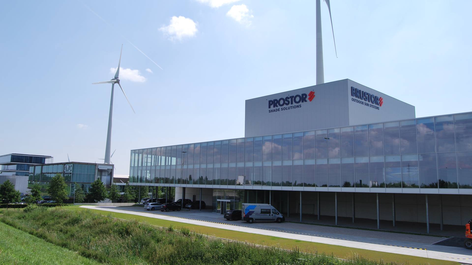Modern kantoorgebouw met glazen gevel en bedrijfslogo, met windturbines op de achtergrond onder een blauwe lucht.