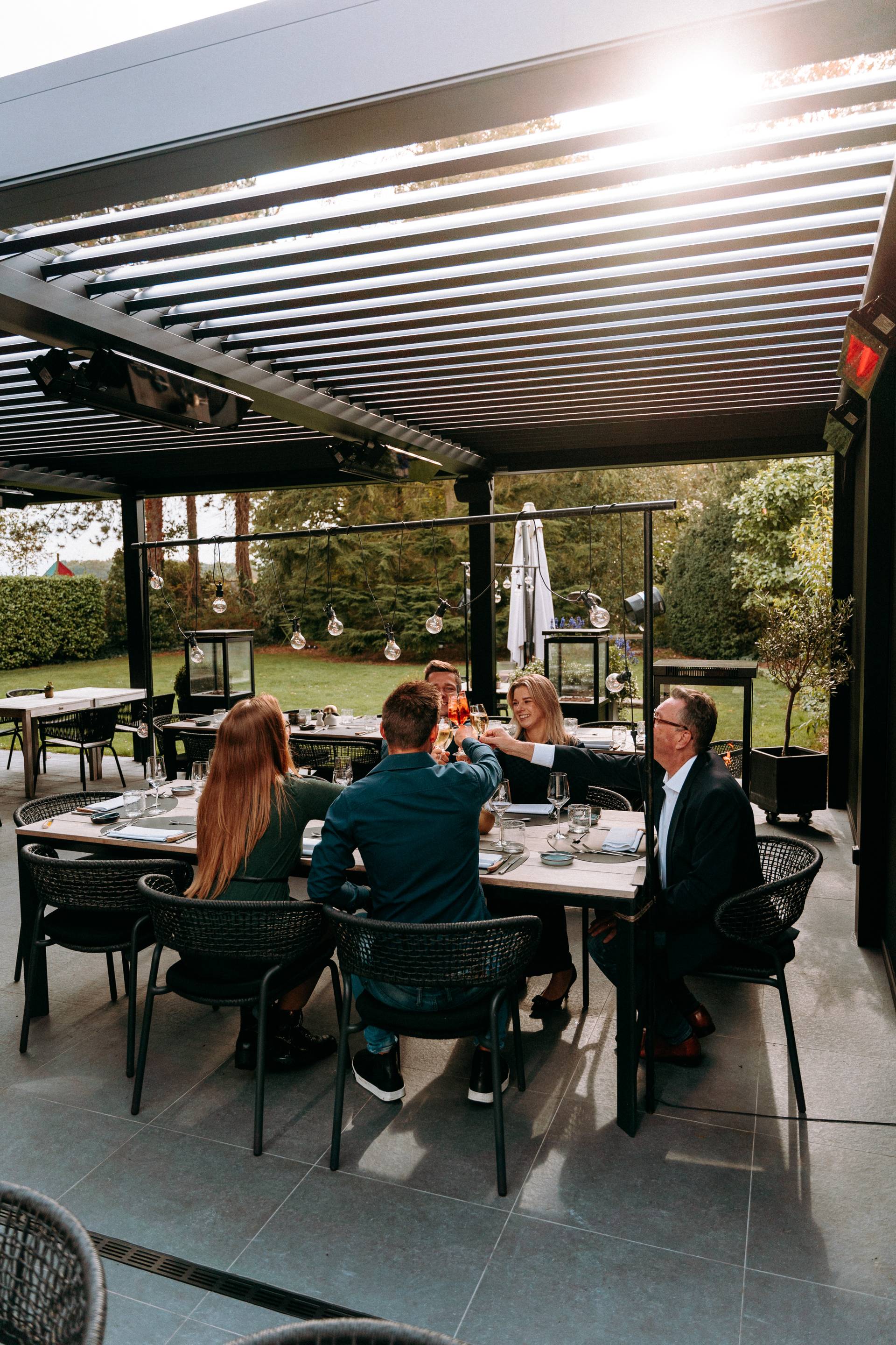 Gruppo che cena all’aperto sotto una pergola moderna con lamelle regolabili e riscaldamento integrato.