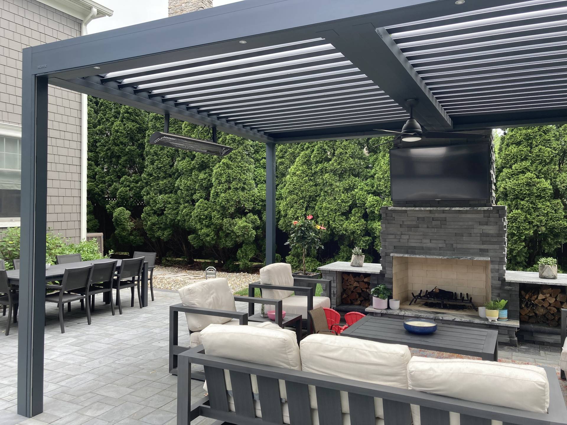 Outdoor seating area beneath an aluminium pergola with tilting louvres, featuring a fireplace, lounge furniture and a landscaped garden backdrop.