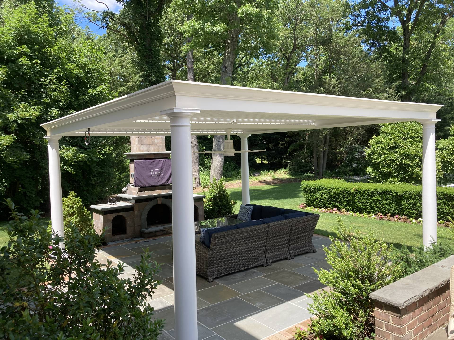 Covered outdoor seating area with a white pergola and columns, featuring wicker furniture and a stone fireplace in a landscaped garden.