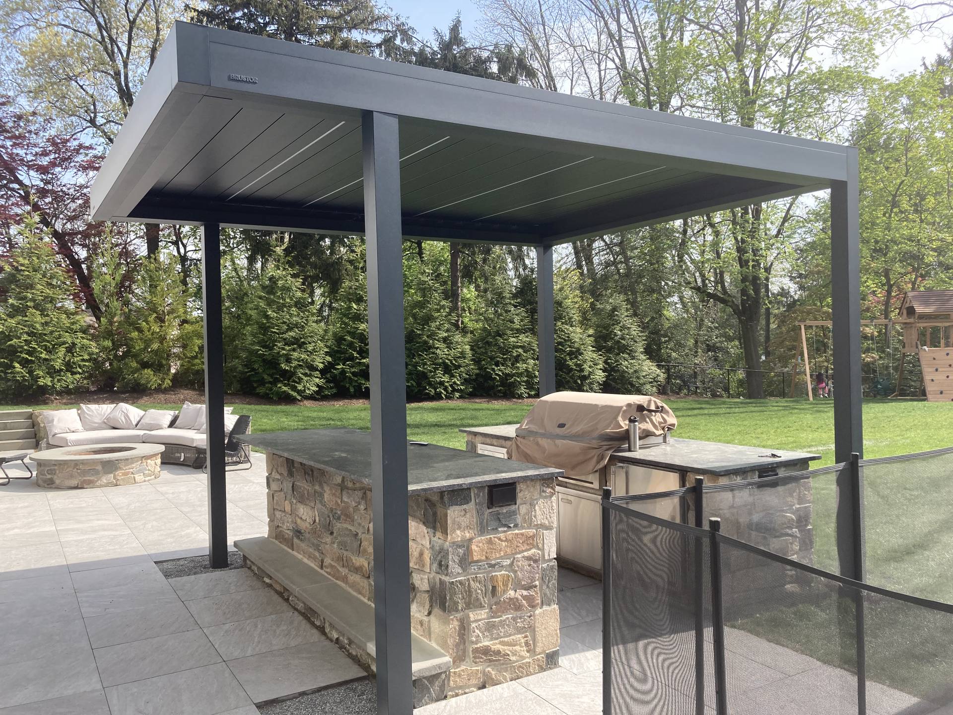 Modern outdoor kitchen area with a stone island and a sleek aluminium pergola, set on a patio overlooking a landscaped lawn and trees.