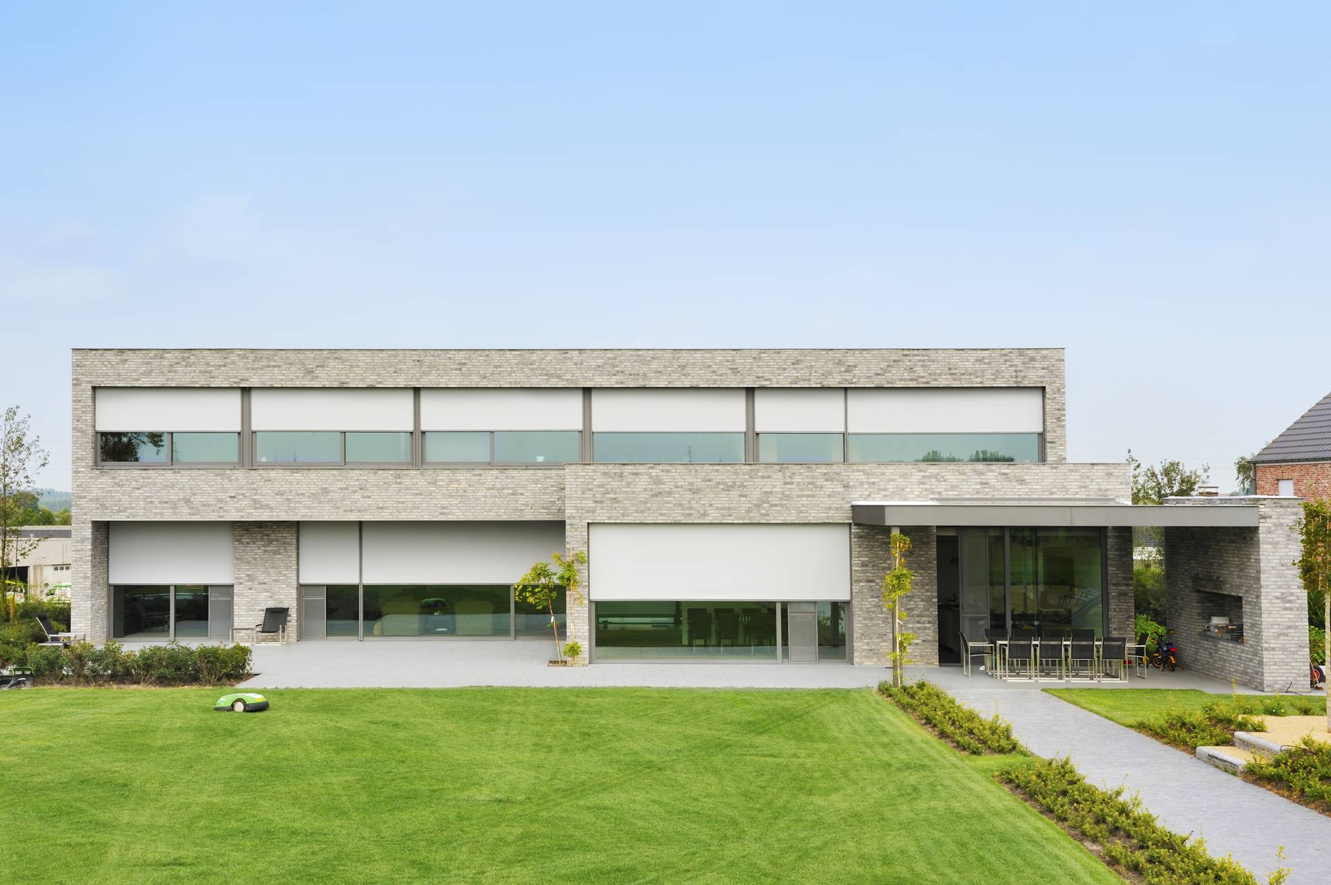 Maison moderne équipée de larges ZIP‑screens en aluminium devant de grandes baies vitrées, donnant sur une pelouse paysagée.