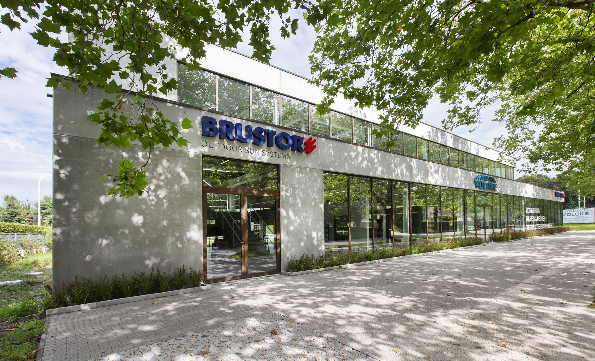 Façade avant du siège moderne de Brustor avec de larges surfaces vitrées, entouré de verdure et ombragé par des arbres