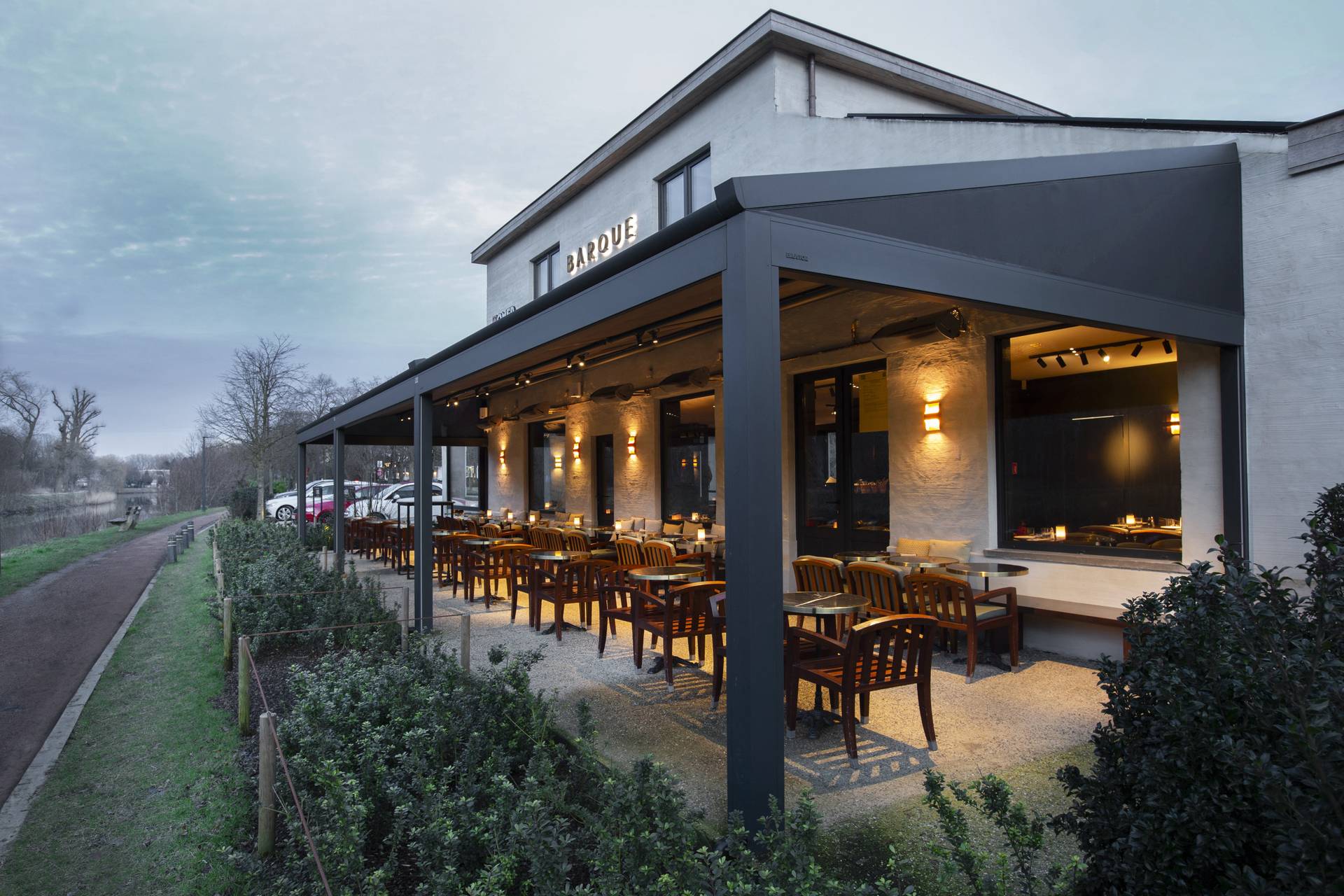 Restaurantterras met een aluminium pergola met geïntegreerde verlichting, die meerdere buiteneettafels voor het gebouw overschaduwt.