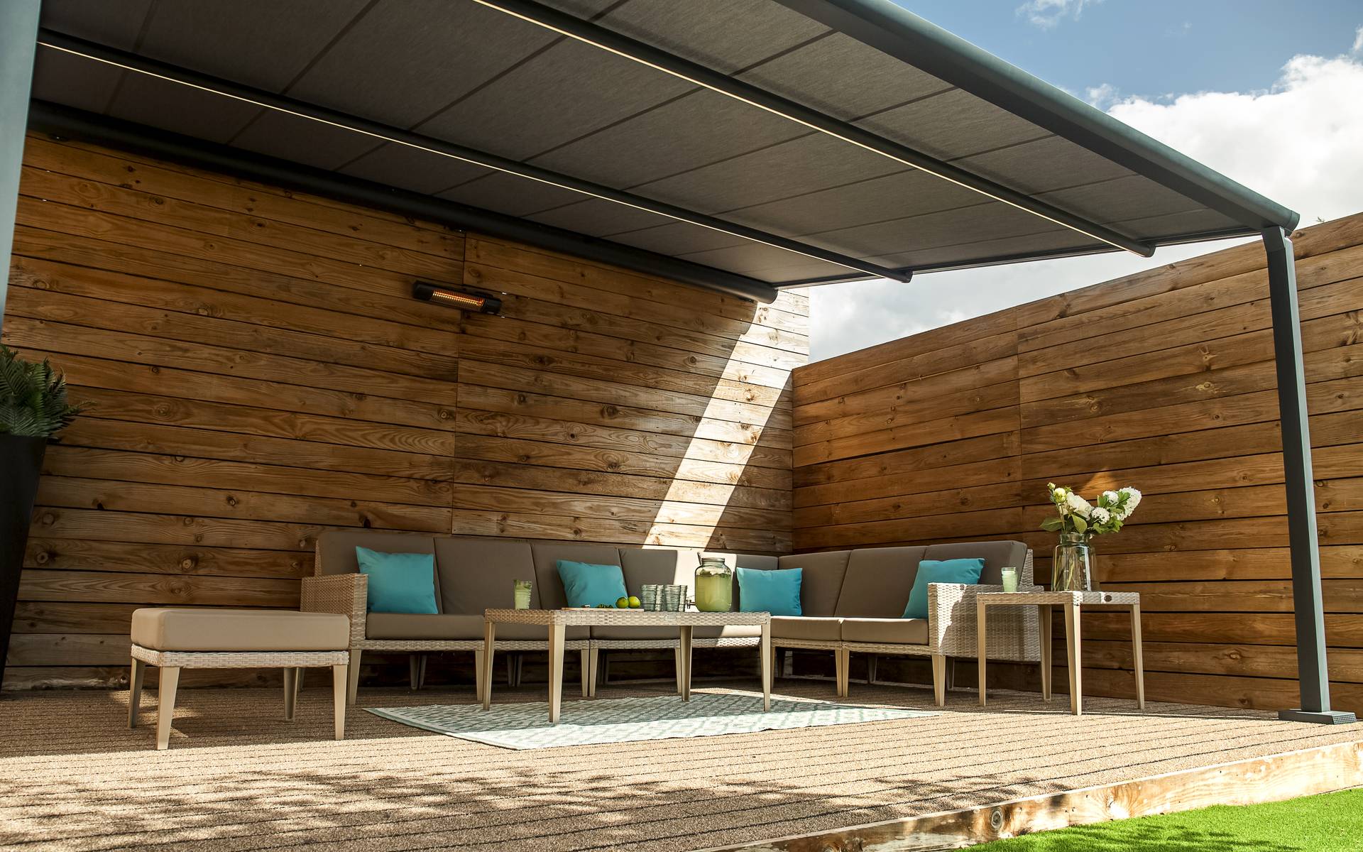 Outdoor lounge area under an aluminium pergola with retractable fabric roof and integrated LED lighting, surrounded by wooden privacy walls.