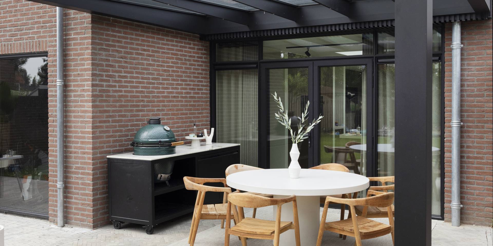 Aluminium patio cover with glass roof attached to a brick house, shading an outdoor dining area with wooden chairs.
