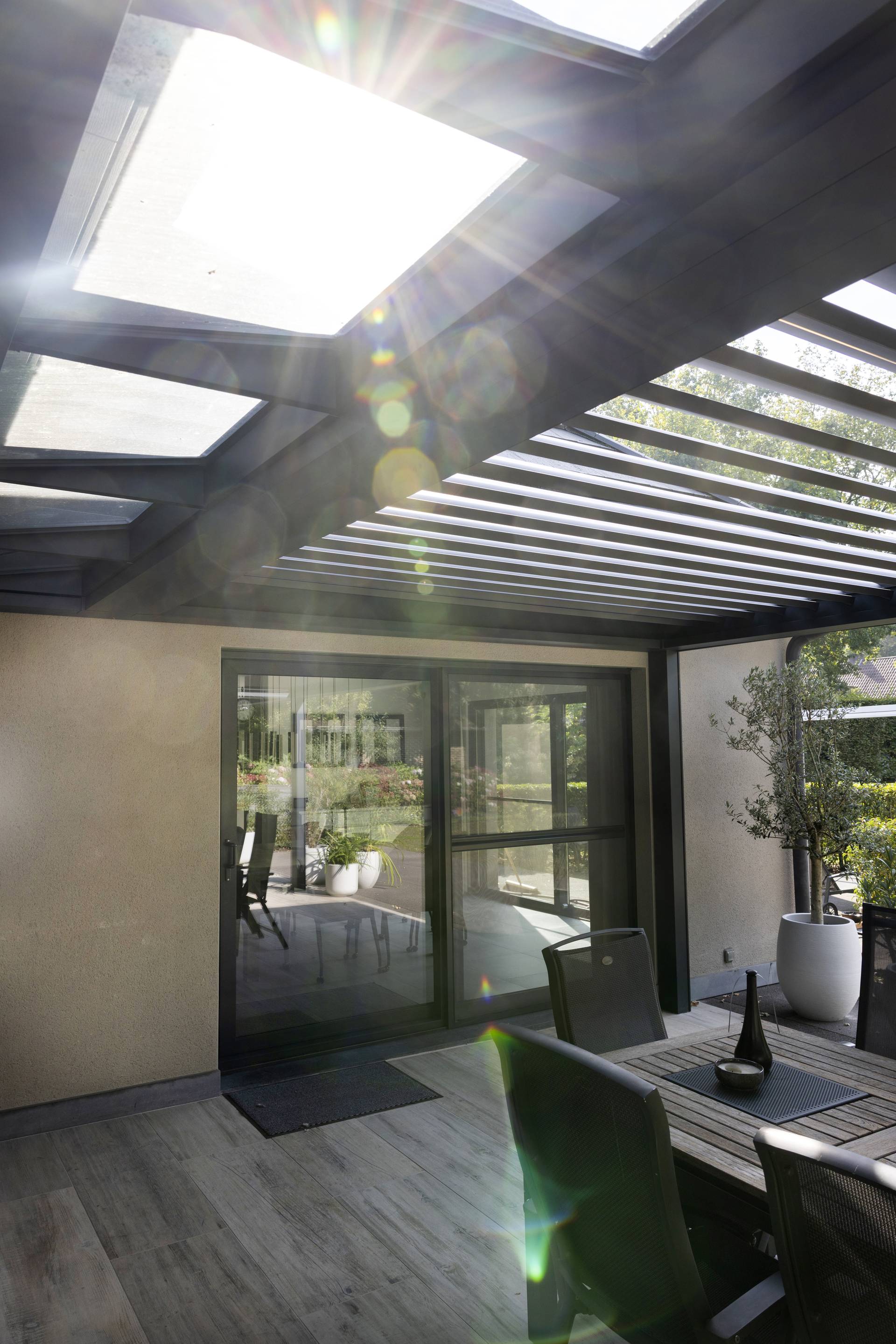 Sonnenlicht, das durch das feste Glaselement und die geöffneten XL‑Lamellen einer Aluminium‑Pergola B720 auf eine moderne Ess‑Terrasse fällt