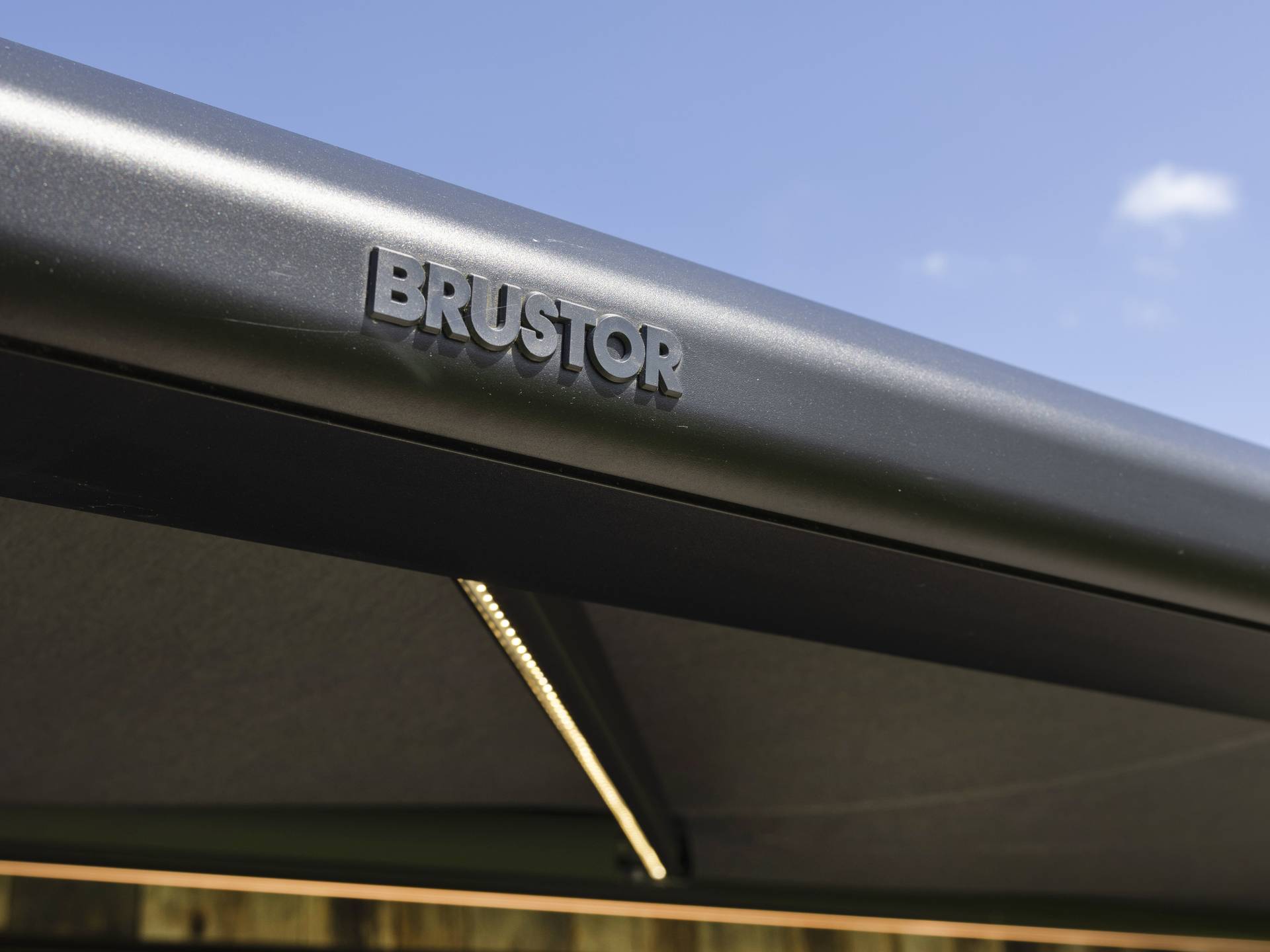 Close-up van een Brustor-zonwering met het logo op het aluminium profiel tegen een blauwe lucht.