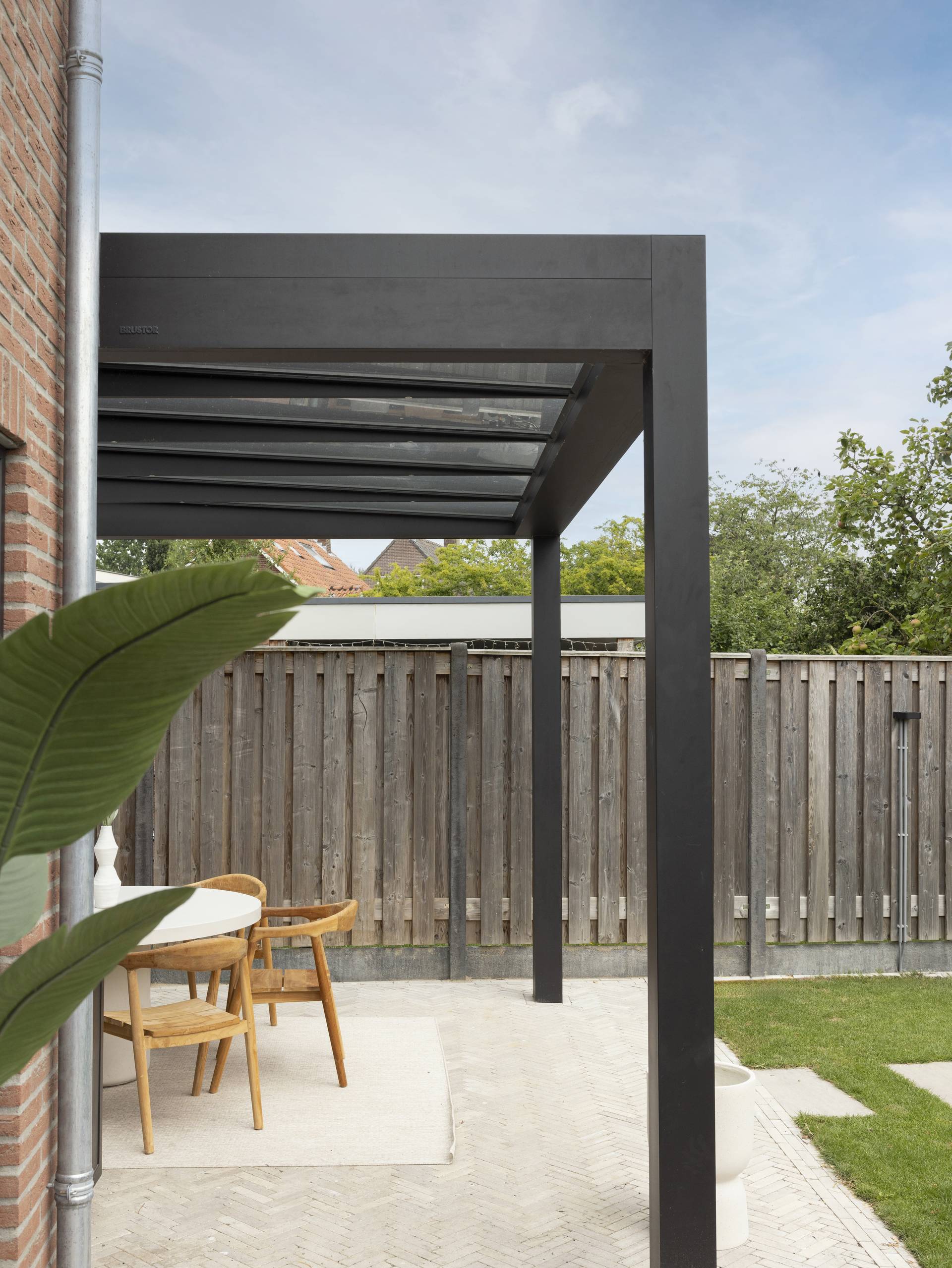 Pergola B800 en aluminium avec toit en verre attenante à la maison, couvrant un espace repas extérieur abrité dans un jardin paisible