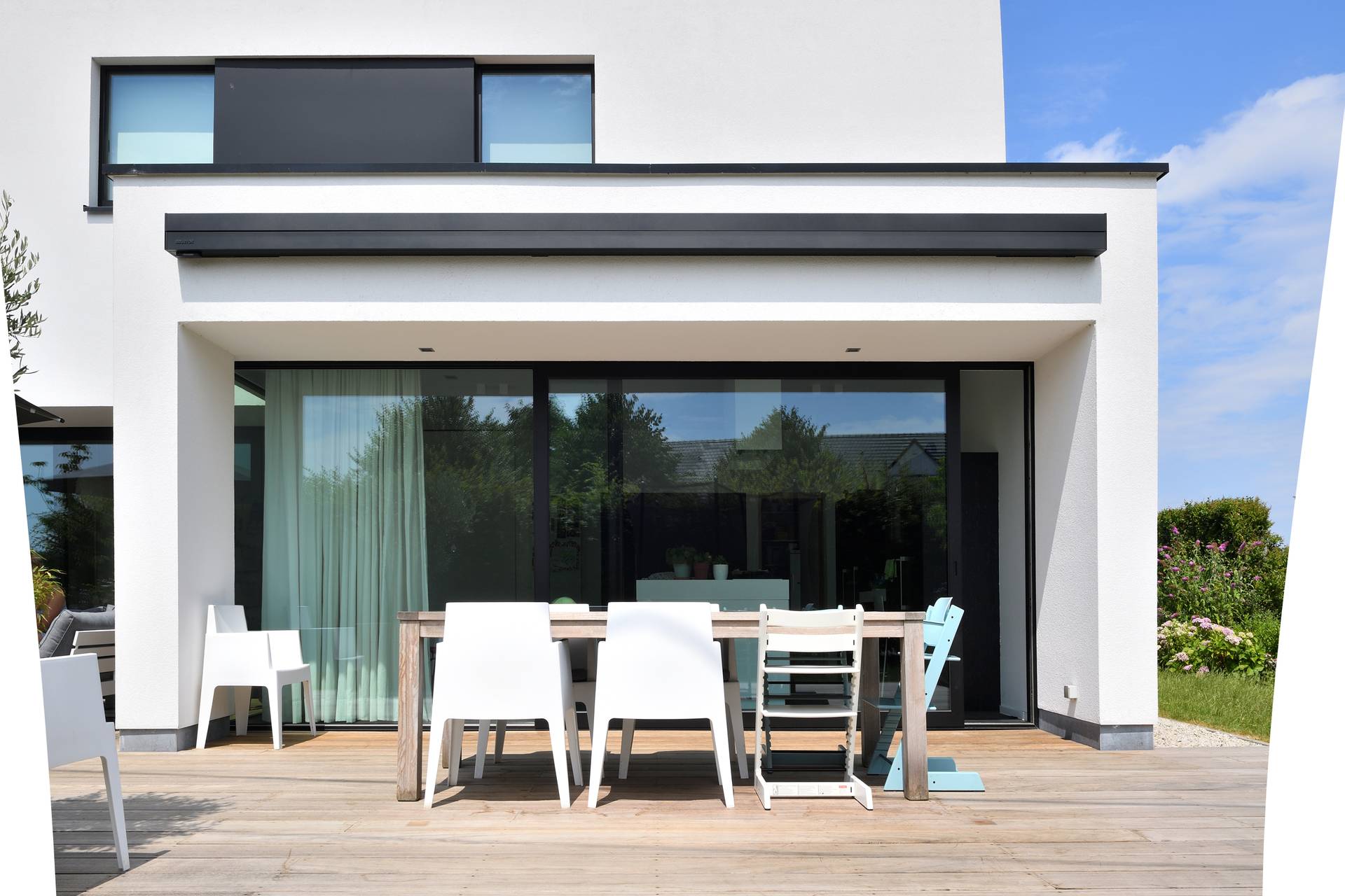 Espace repas extérieur avec table en bois et chaises blanches sur une terrasse attenante à une maison moderne aux grandes baies vitrées coulissantes.