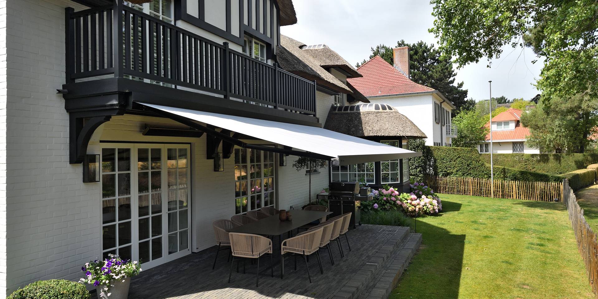 Aluminium awning providing shade over a terrace dining area on a traditional house façade, overlooking a landscaped garden.