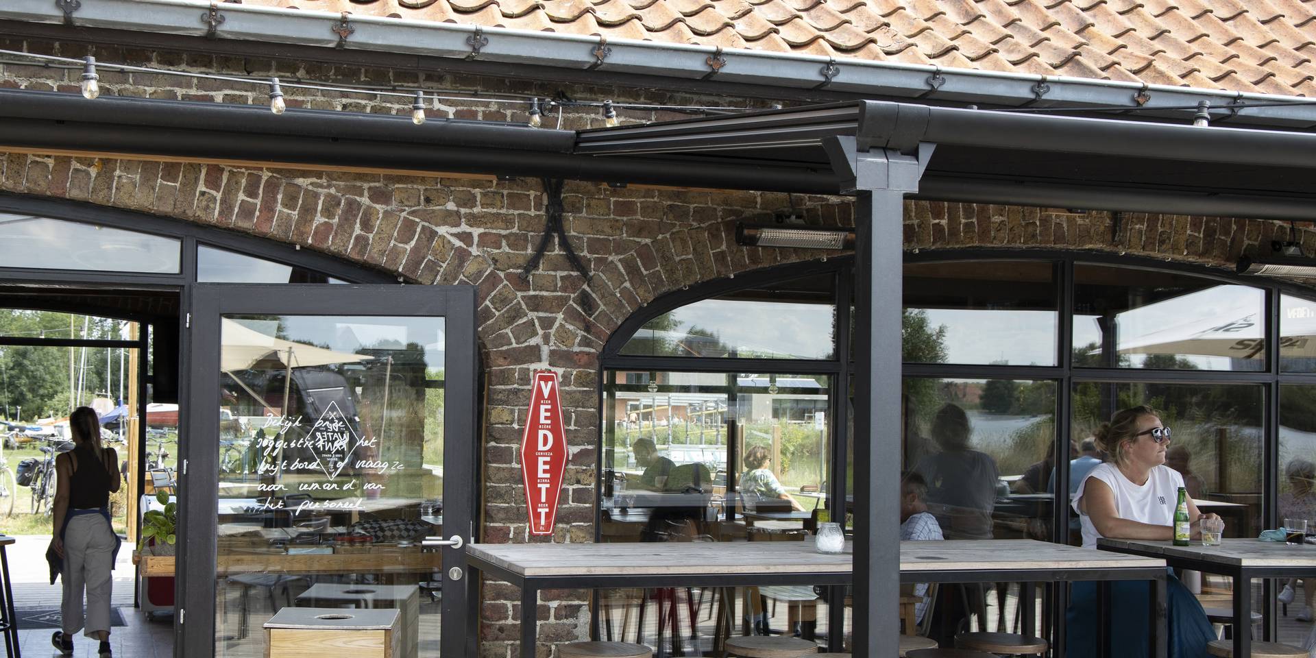 Außenbereich eines Gastronomiebetriebs mit einer Brustor‑Pergola aus Aluminium und ausfahrbarem Stoffdach, eingebettet in eine rustikale Backsteinfassade für angenehmen Schatten.