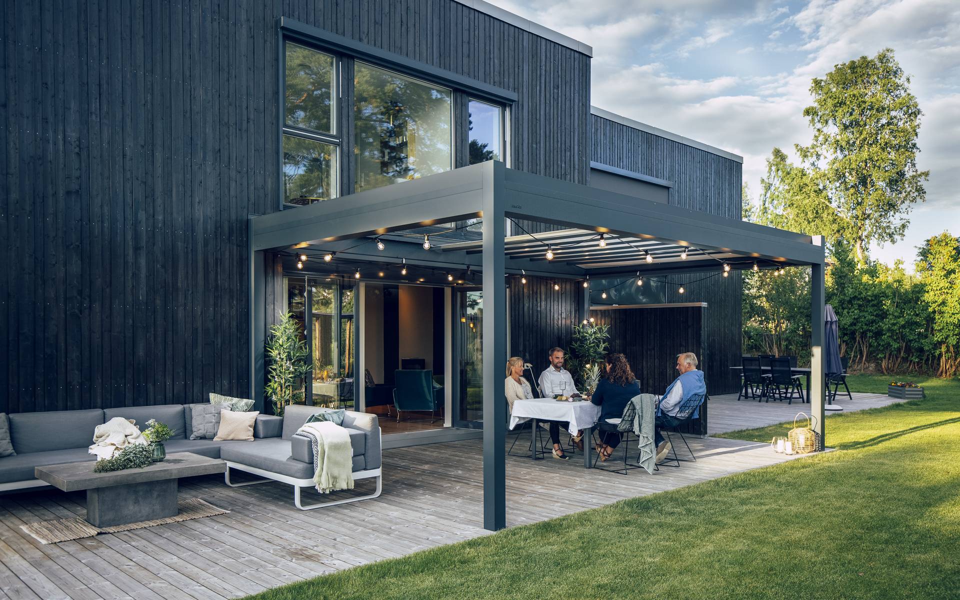 Pergola en aluminium à lames orientables couvrant un espace repas et lounge extérieur sur une terrasse en bois attenante à une maison moderne.