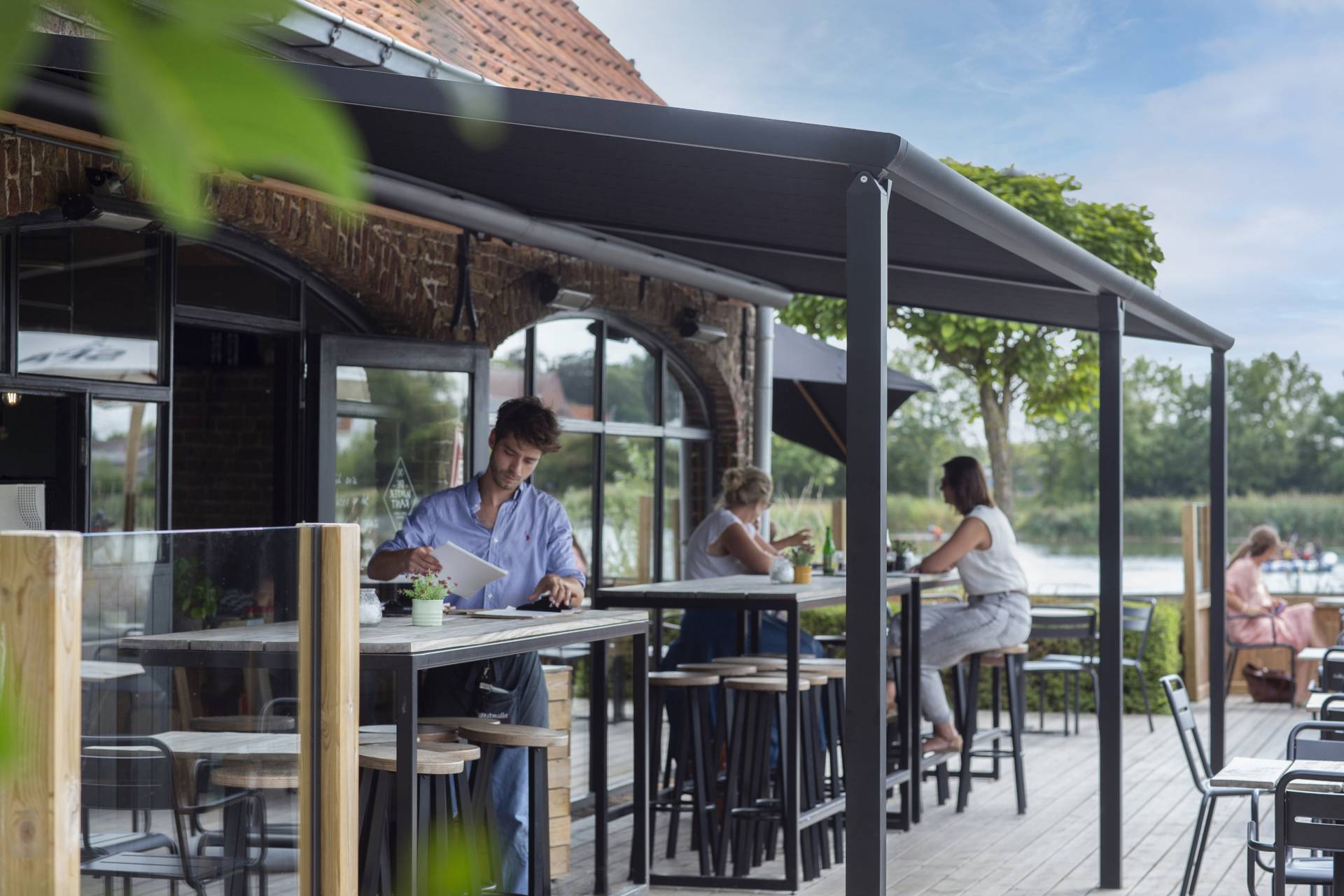 Buitenterras van een horecazaak met een aluminium Brustor‑pergola met oprolbaar doekdak, die schaduw en comfort biedt in een sfeervolle locatie aan het water.