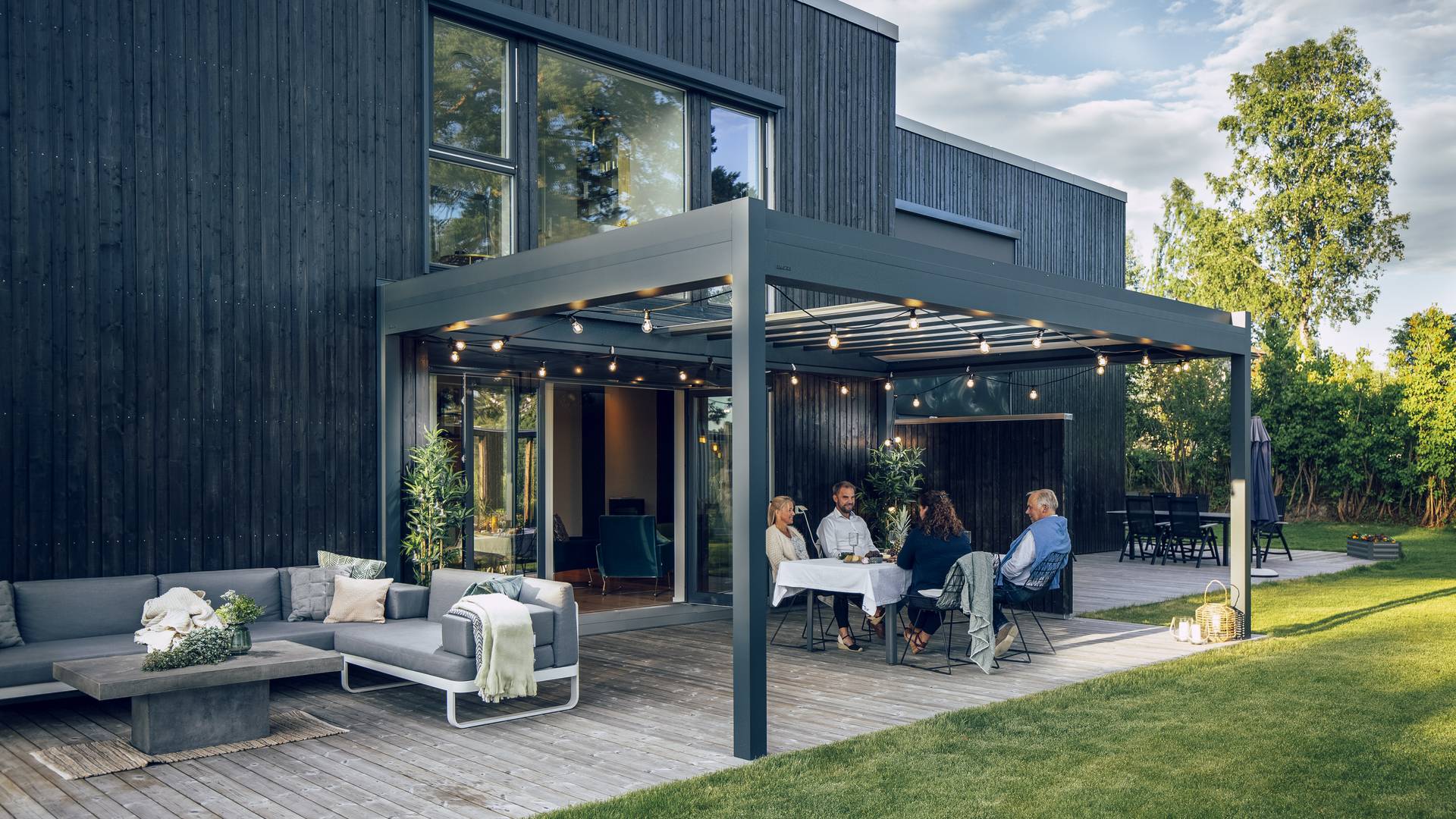 Pergola en aluminium avec toit rétractable couvrant un espace repas et lounge extérieur sur une terrasse en bois attenante à une maison moderne.