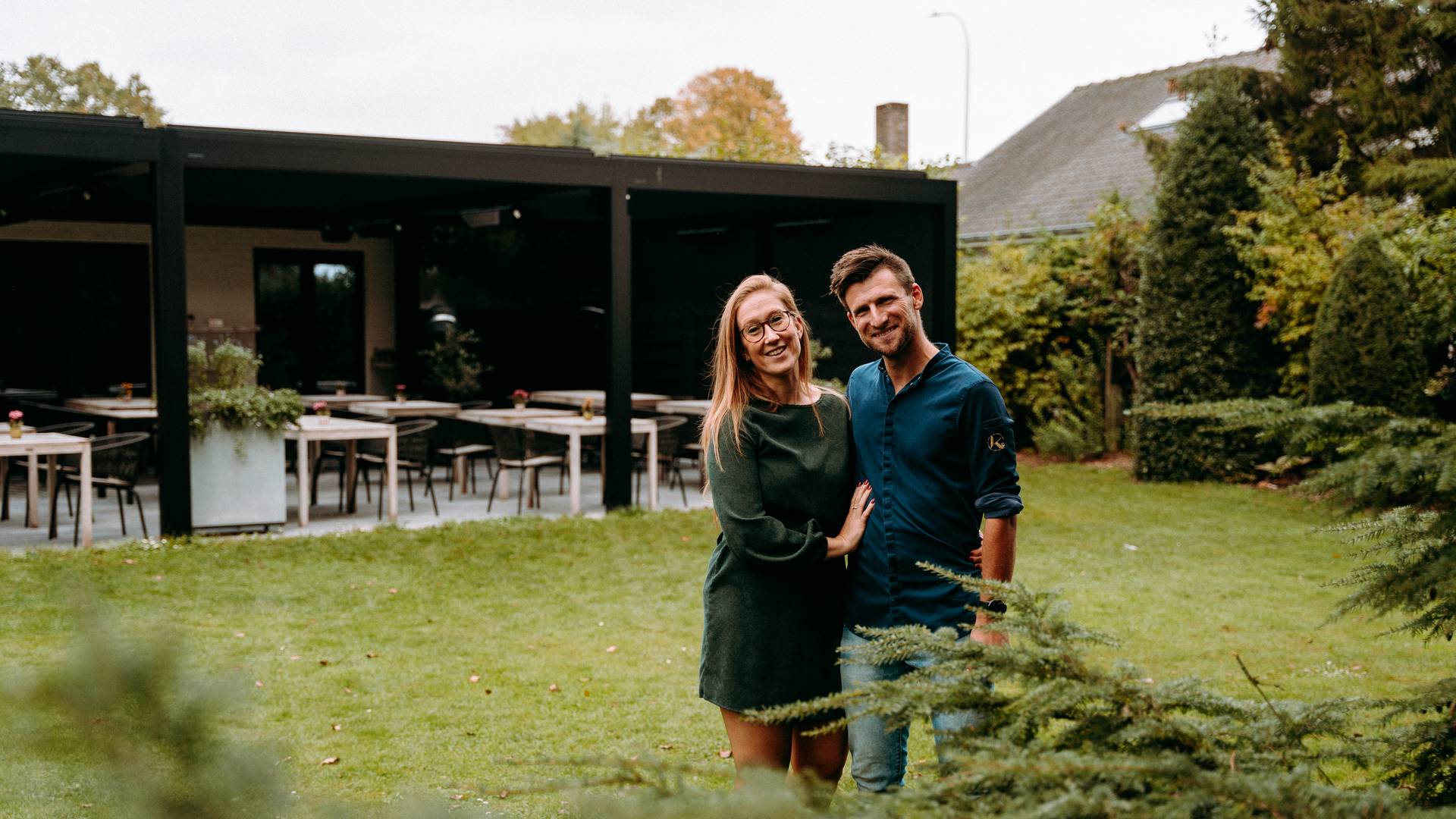 Coppia in piedi in un’area verde con una pergola moderna e zona pranzo esterna sullo sfondo.