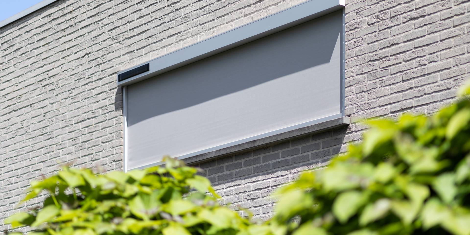 Aluminium ZIP‑screen op zonne‑energie aan een lichtgekleurde bakstenen gevel, met geïntegreerd zonnepaneel boven het doek.