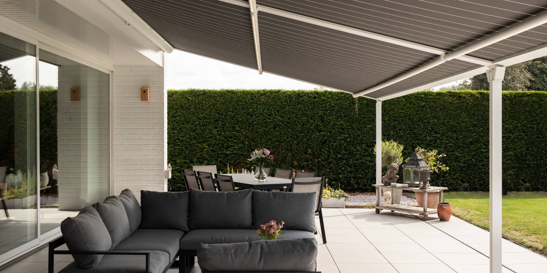 Store banne rétractable entièrement déployé au‑dessus d’une terrasse avec espace lounge et repas extérieur, attenant à une maison blanche.
