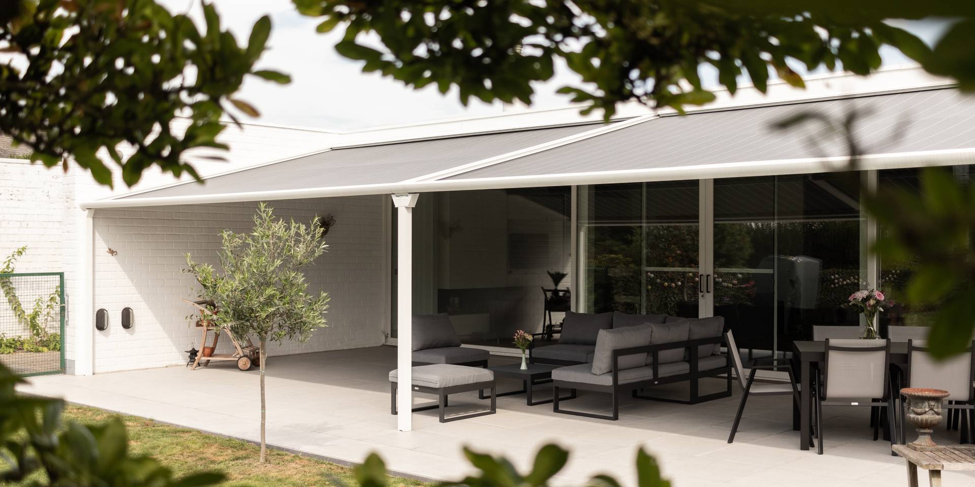 Aluminium pergola‑zonwering met uitschuifbaar doekdak aan een moderne woning, die schaduw biedt boven een buitenlounge en eethoek met zicht op de tuin.
