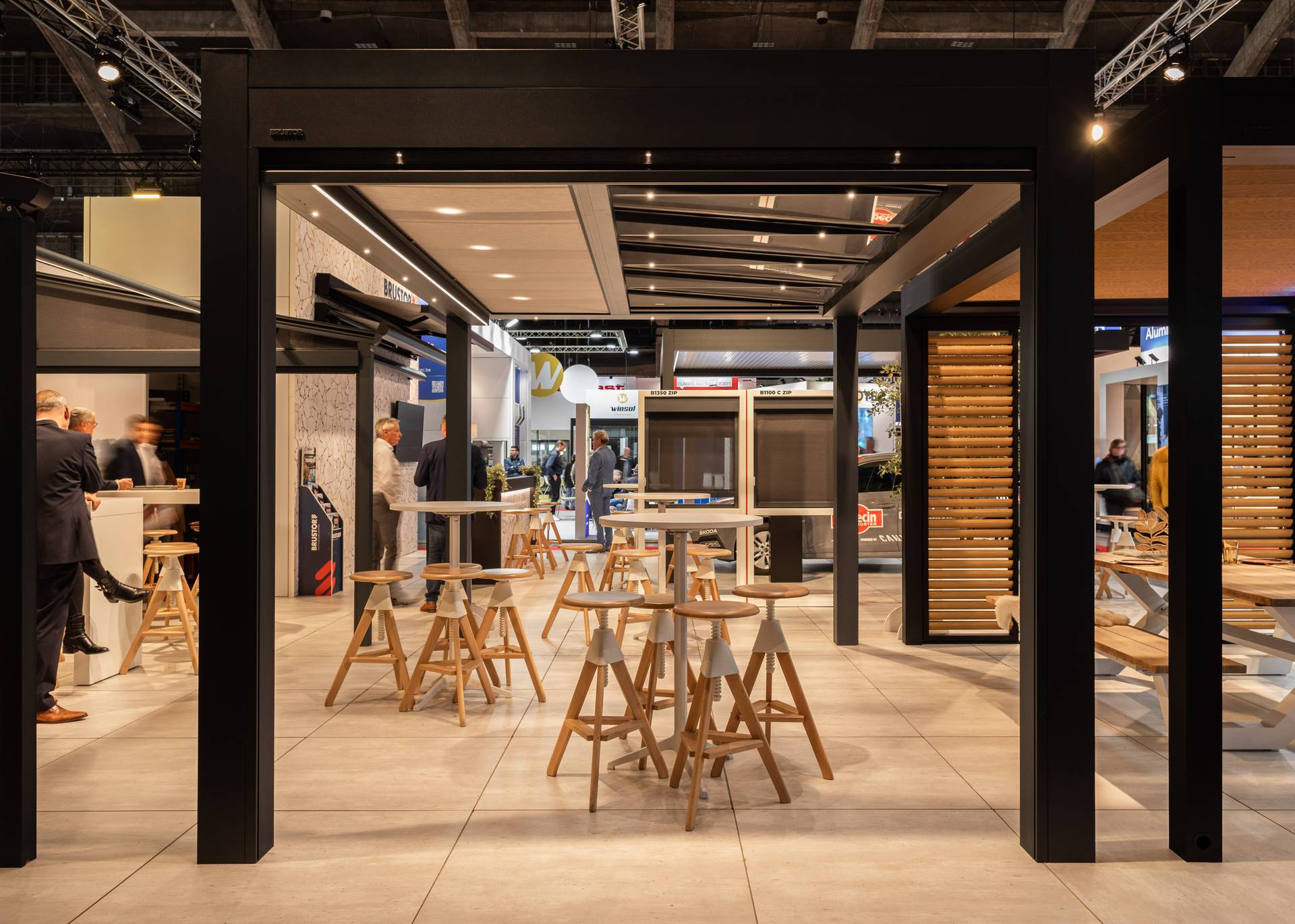 Beursruimte met een aluminium pergola met geïntegreerde led‑verlichting, hoge tafels en krukken, en productpresentaties op de achtergrond.