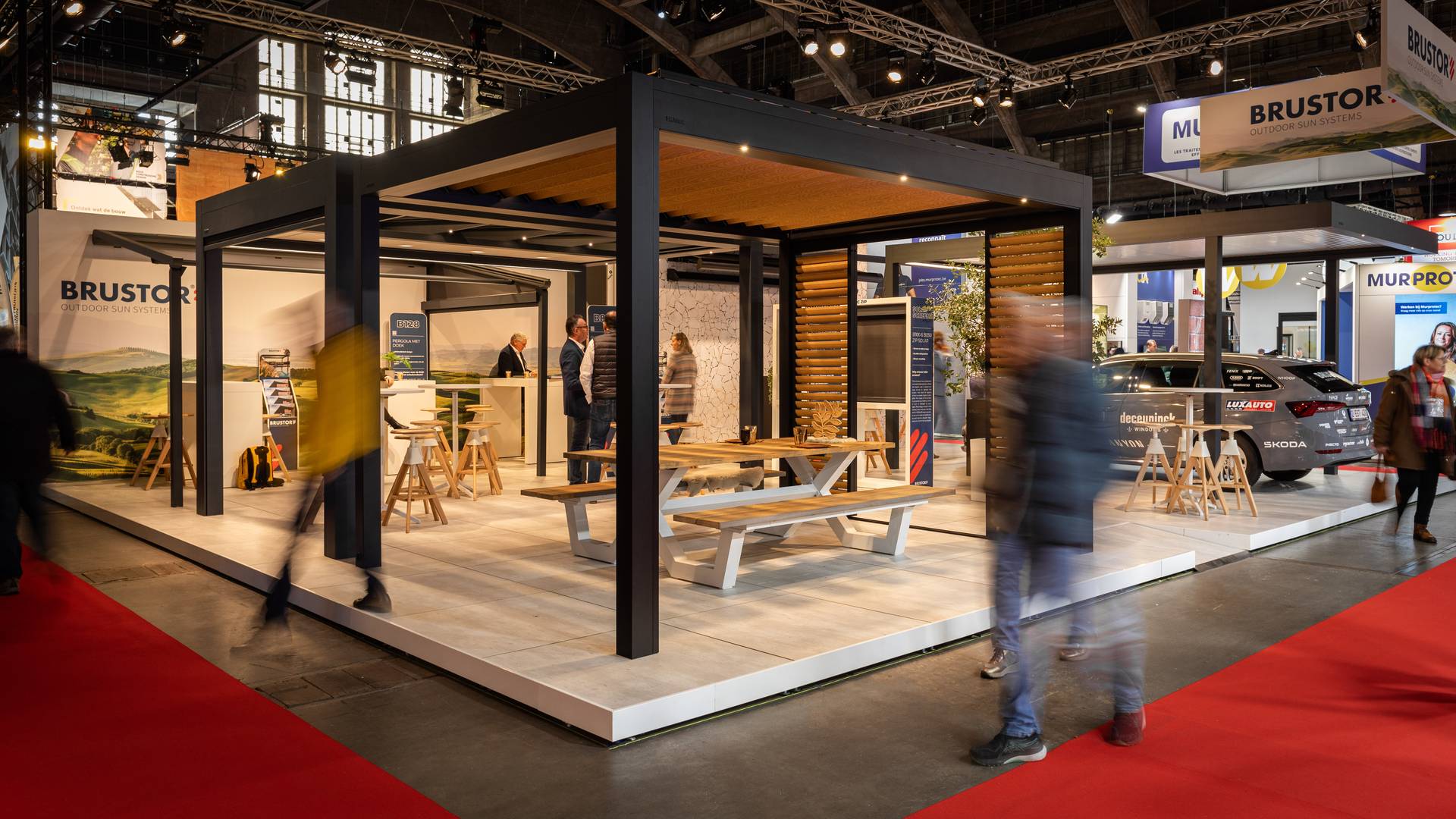 Stand Brustor au salon Batibouw avec pergola en aluminium éclairée, mobilier extérieur et présentations de produits, tandis que des visiteurs passent.
