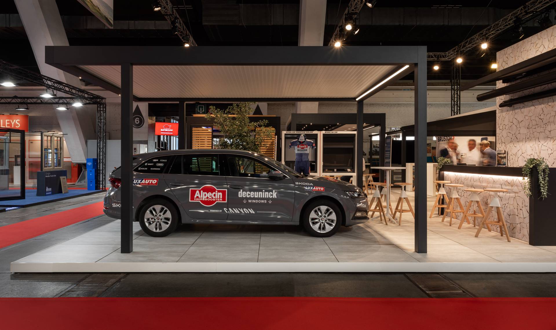 Alpecin–Deceuninck‑teamwagen naast een aluminium pergola op de Brustor‑stand, met outdoor opstellingen op de achtergrond.