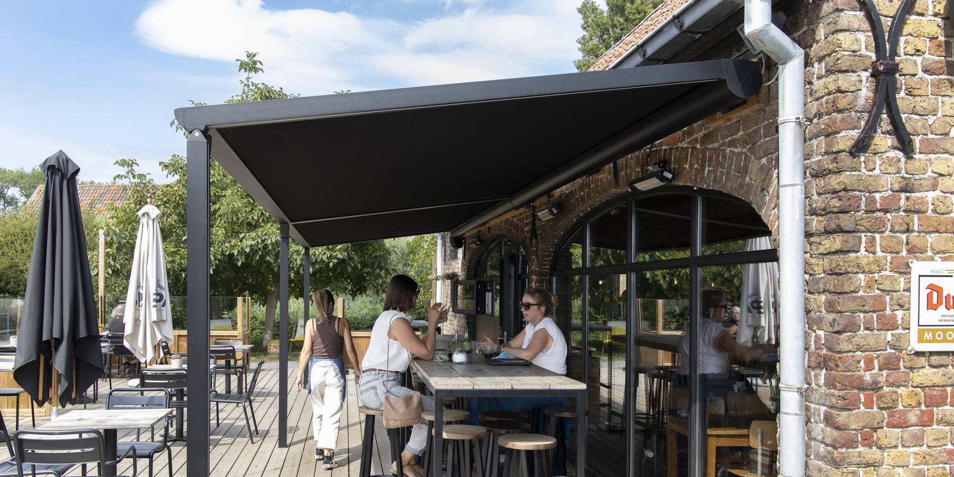 Außenterrasse eines Gastronomiebetriebs mit einer Brustor‑Pergola aus Aluminium und ausfahrbarem Stoffdach, die Schatten und Komfort in moderner Umgebung bietet.