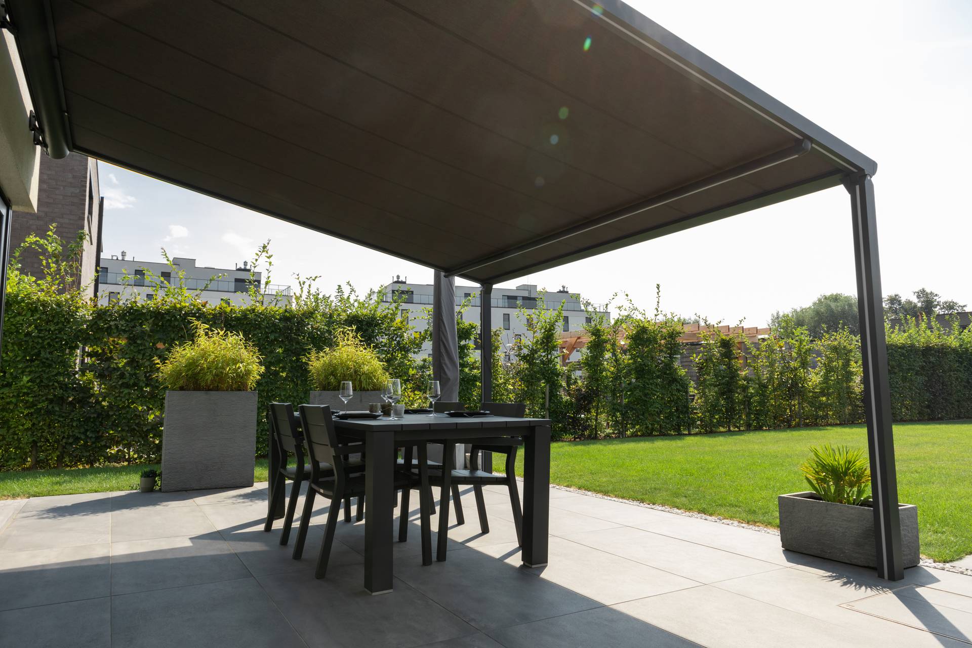 Uitschuifbaar zonnescherm dat schaduw biedt boven een buiteneettafel op een betegeld terras met uitzicht op een groene tuin.