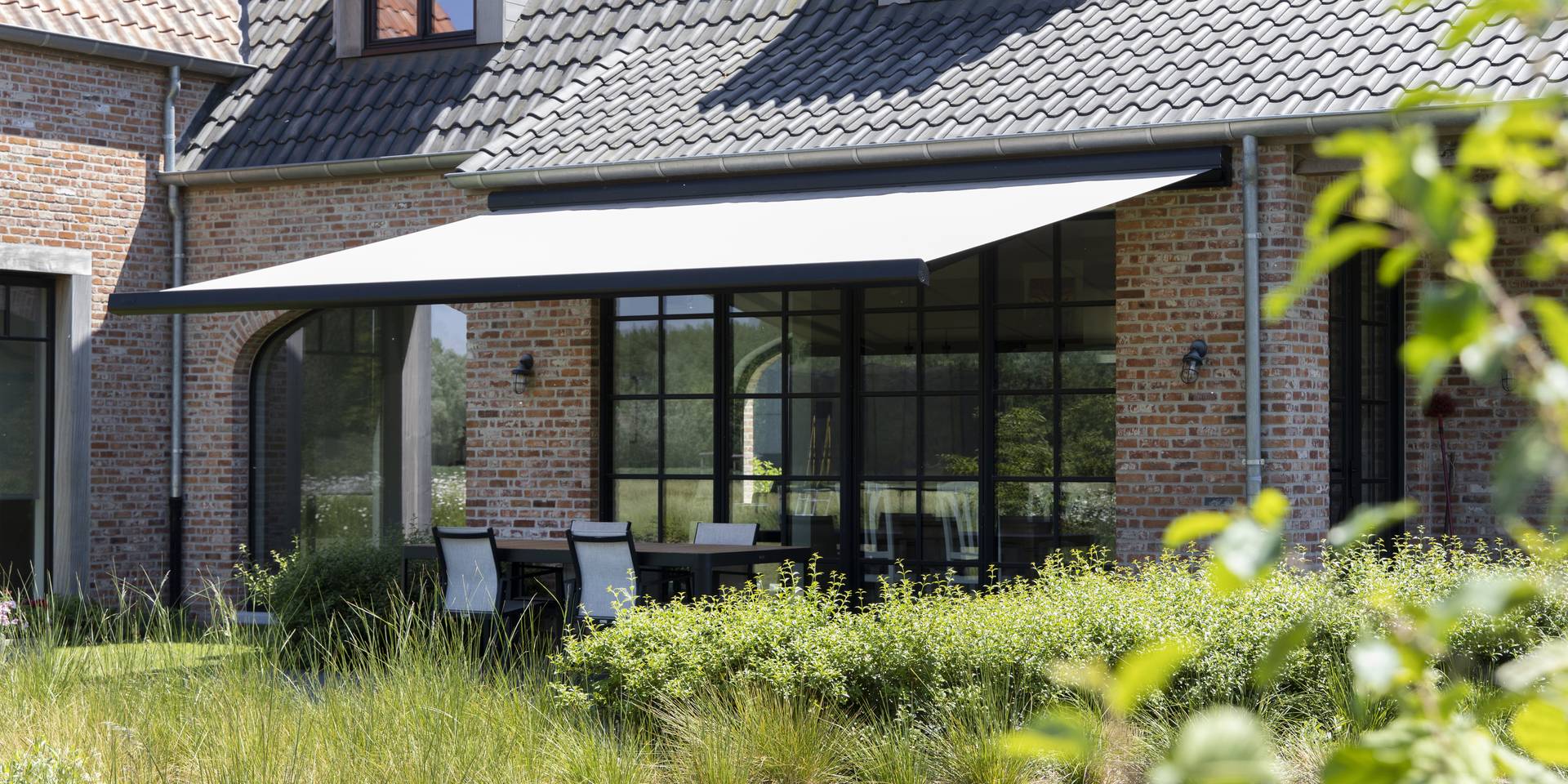 Aluminium awning extended over a terrace at a traditional brick house, shading an outdoor dining area surrounded by garden greenery.