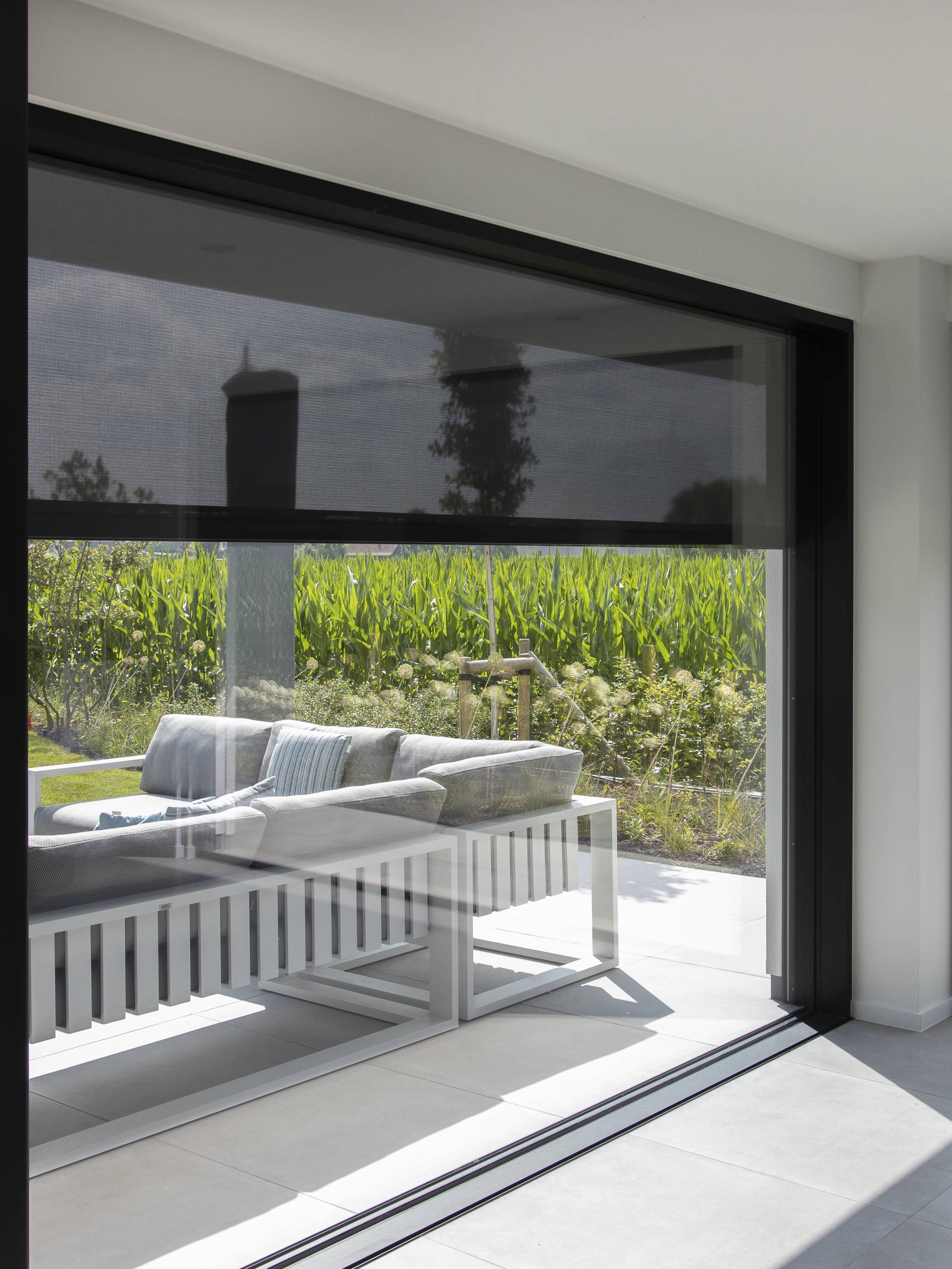 Vue intérieure de grandes fenêtres d’angle avec stores screen extérieurs partiellement abaissés, filtrant la lumière et donnant sur une terrasse de jardin.
