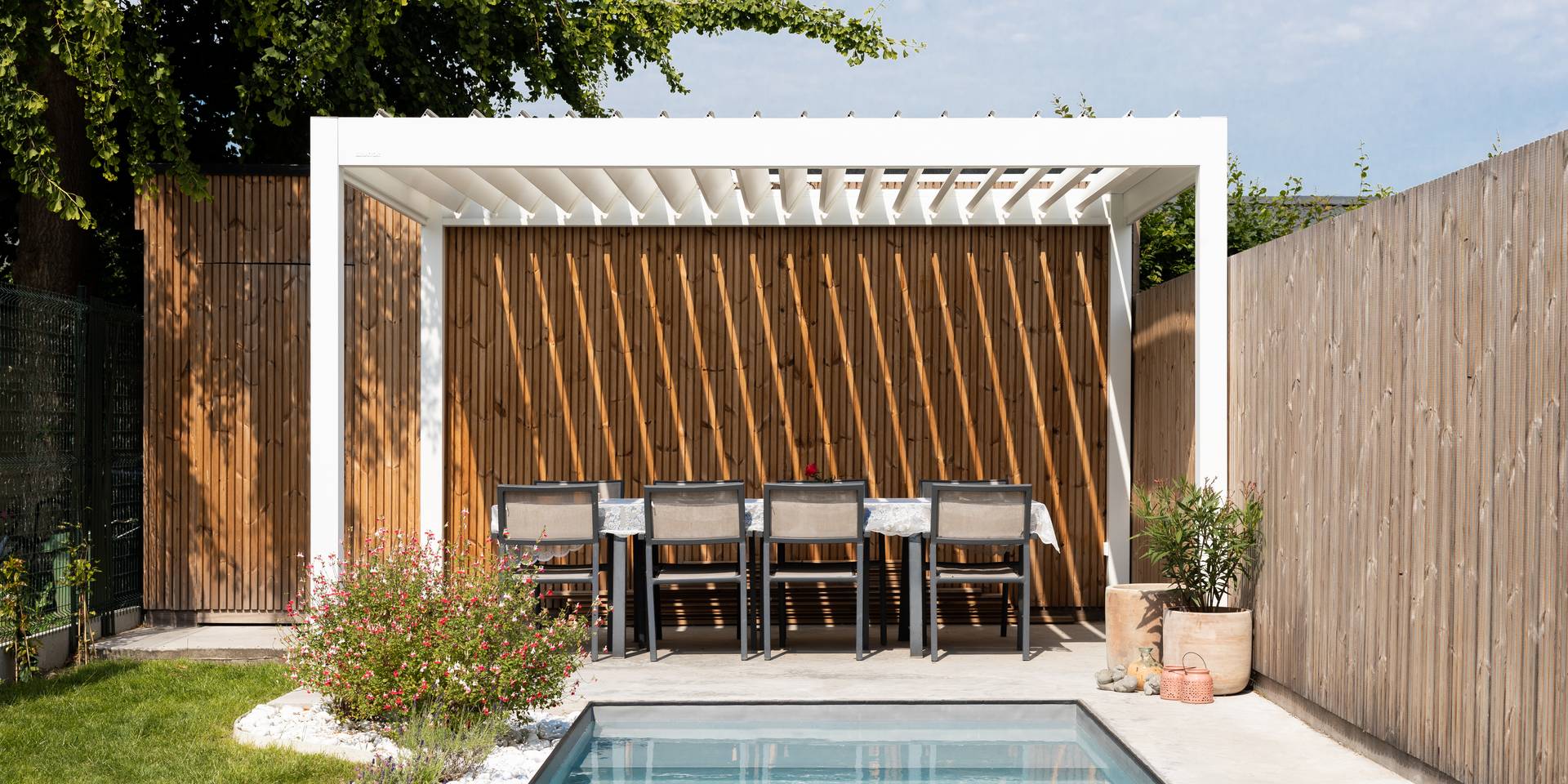 Aluminium pergola with tilting louvred roof and wooden slat wall shading an outdoor dining area beside a pool in a landscaped garden.