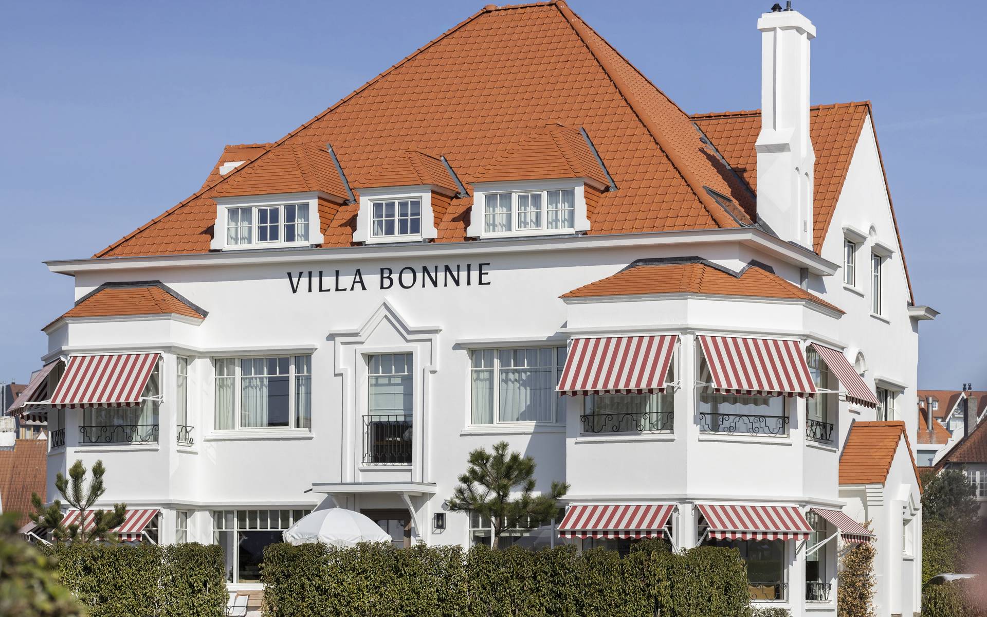 Frontansicht der Villa Bonnie mit rot‑weiß gestreiften Brustor‑Ausfallmarkisen, die den Küstencharakter betonen und stilvollen Sonnenschutz bieten.