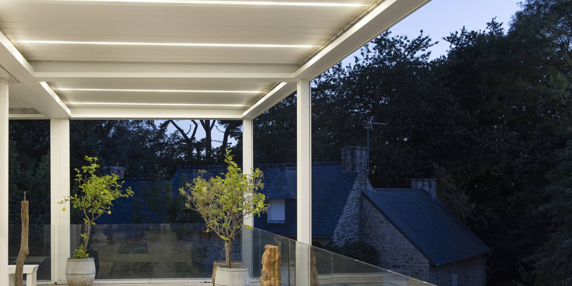 Aluminium pergola with integrated LED lighting above a terrace with glass balustrade, overlooking surrounding trees and rooftops at dusk.