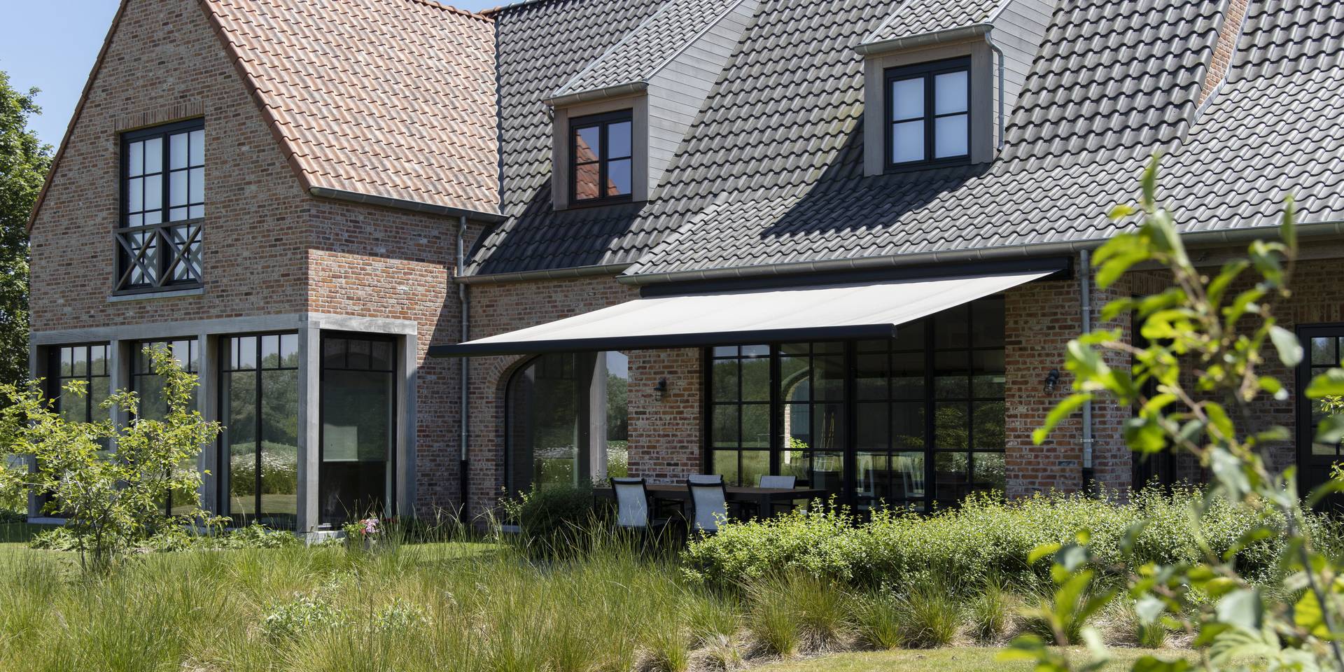 Store banne en aluminium offrant de l’ombre à une terrasse d’une maison en briques, avec vue sur un jardin paysager.