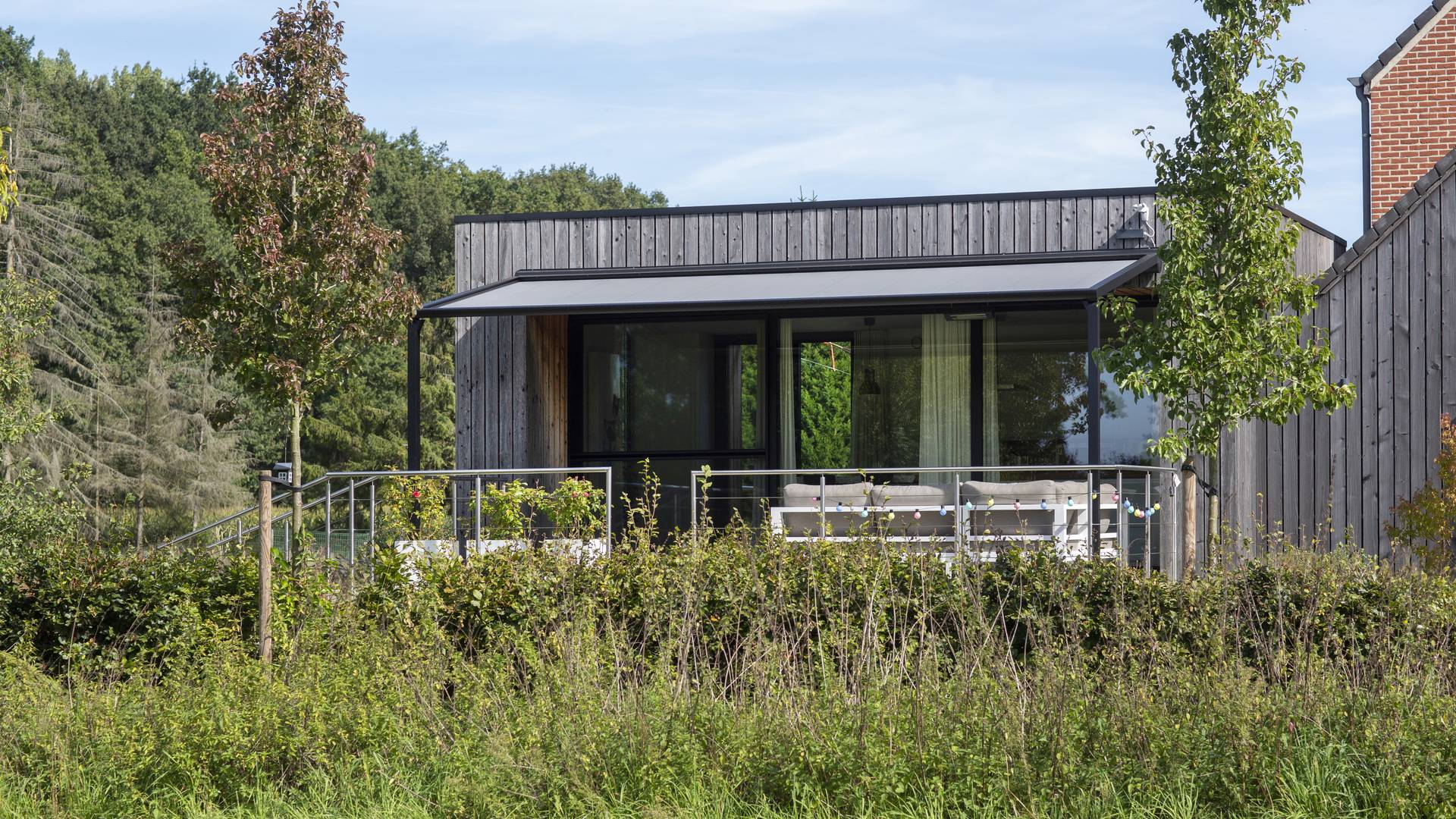 Aluminium pergola met oprolbaar doekdak aan een moderne houten woning met schaduwrijk terras en loungehoek in groene tuin