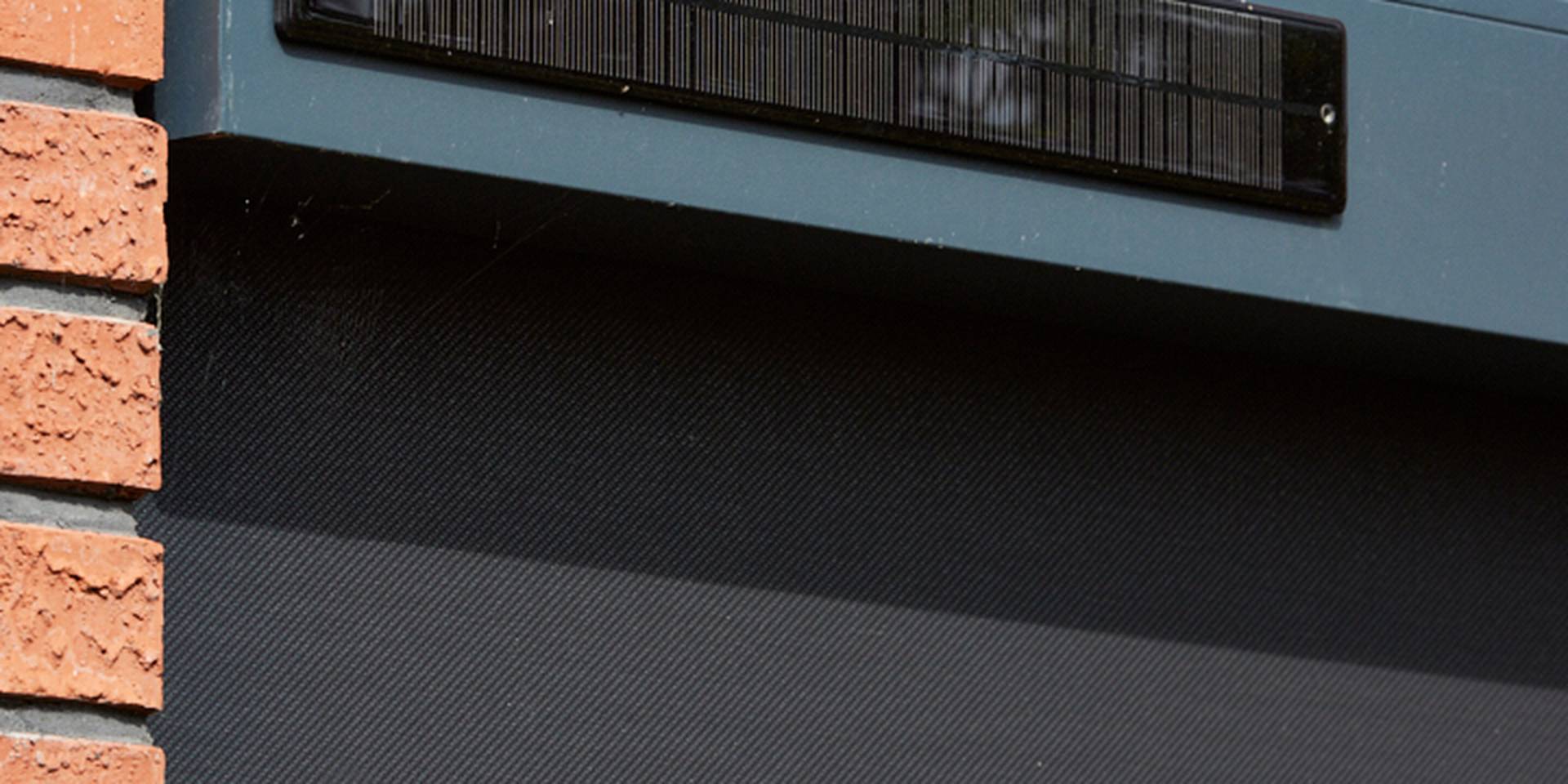 Close‑up van een aluminium ZIP‑screen op zonne‑energie met geïntegreerd zonnepaneel boven het doek, gemonteerd aan een bakstenen gevel.