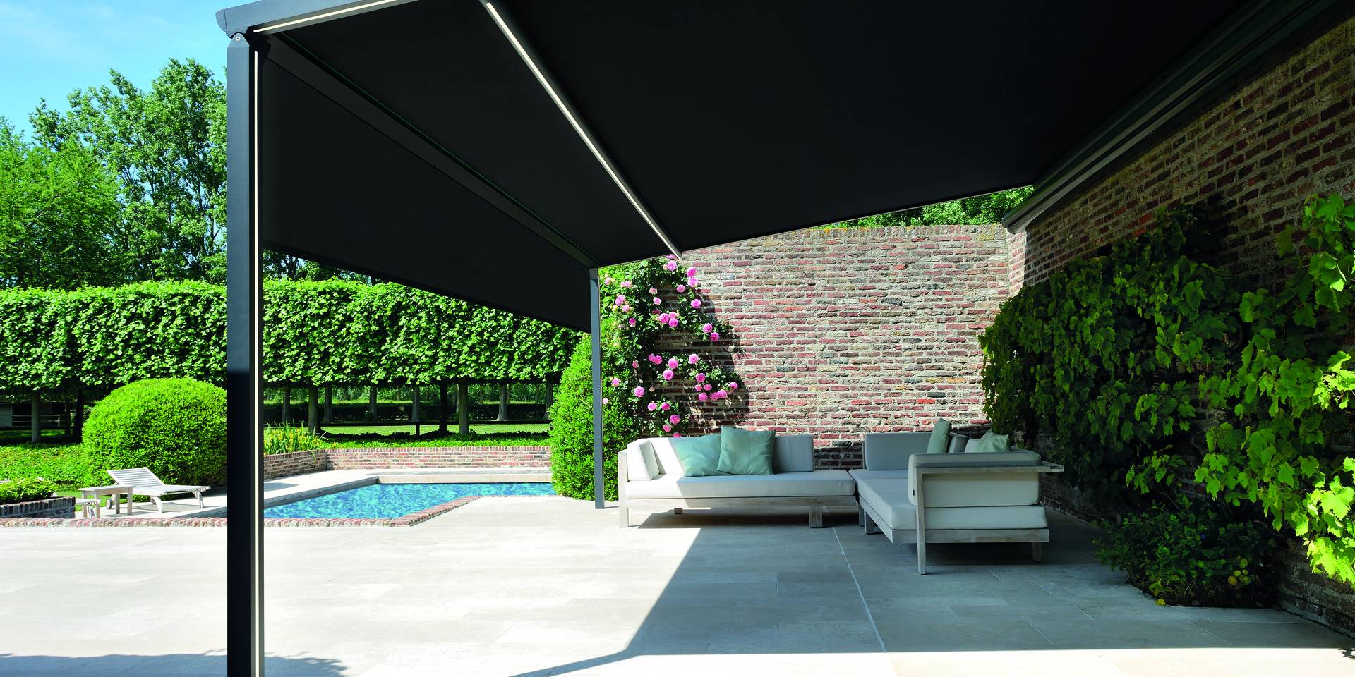 Aluminium pergola‑zonwering met uitschuifbaar doekdak dat schaduw biedt boven een terraslounge naast een zwembad en bakstenen tuinmuur.