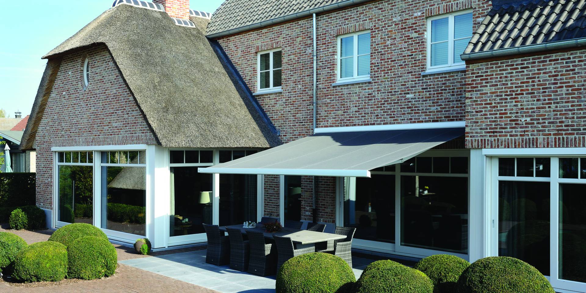 Aluminium awning extended over a terrace dining area at a traditional brick house, providing shade along the rear façade.