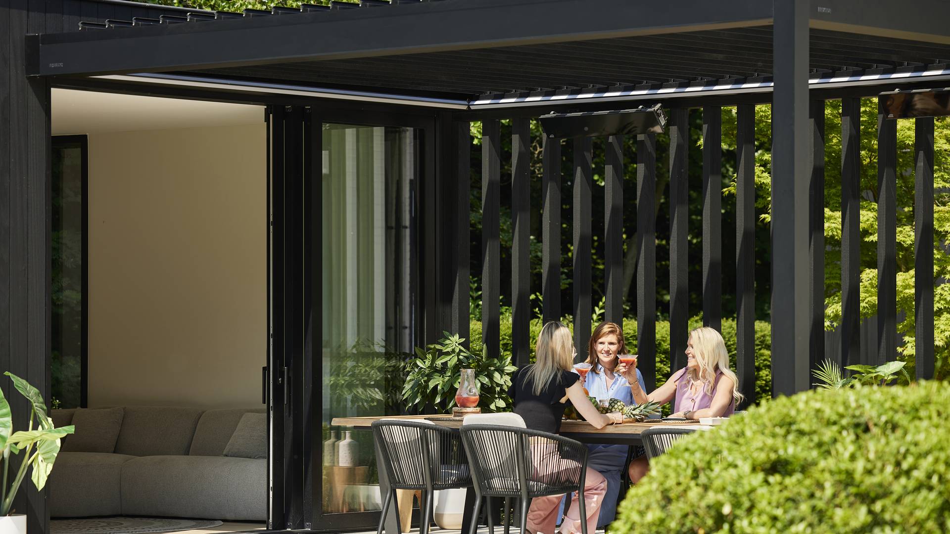 Outdoor dining area under a modern aluminium pergola with vertical slats, connected to the house and surrounded by garden greenery.