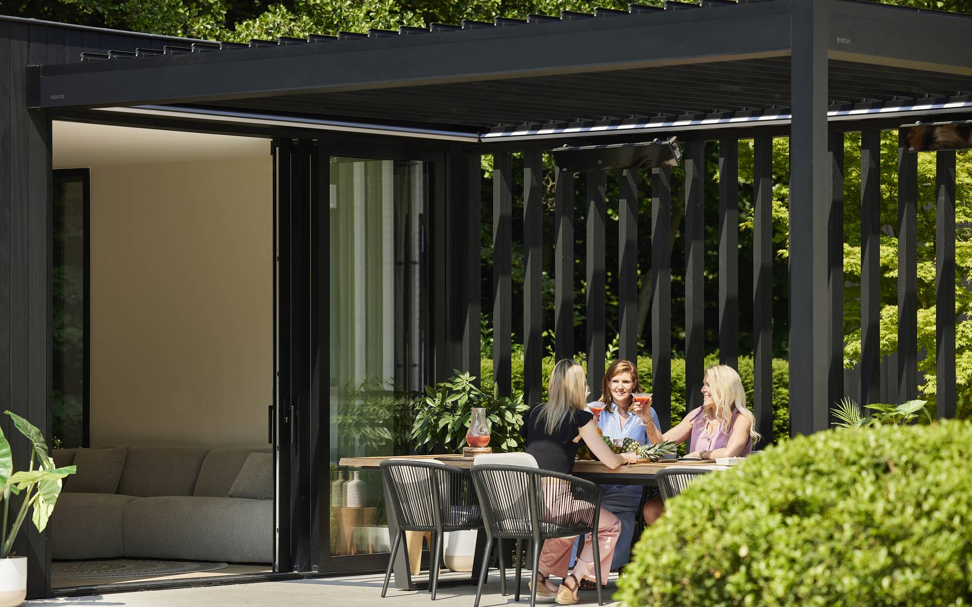 Outdoor dining area under a modern aluminium pergola with vertical slats, connected to the house and surrounded by garden greenery.