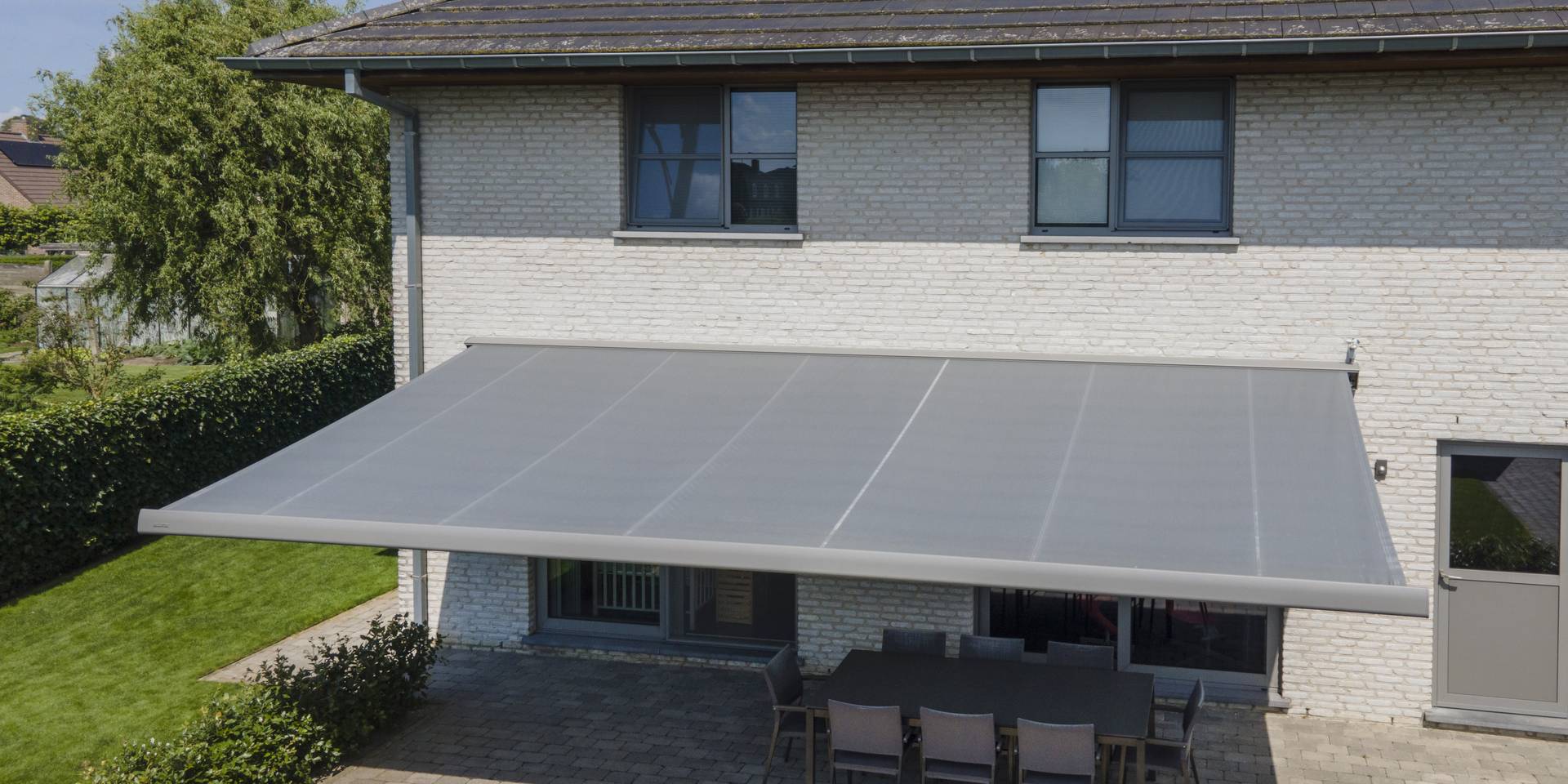Grand store banne rétractable déployé au‑dessus d’une vaste terrasse d’une maison moderne en briques, ombrageant l’espace repas extérieur.