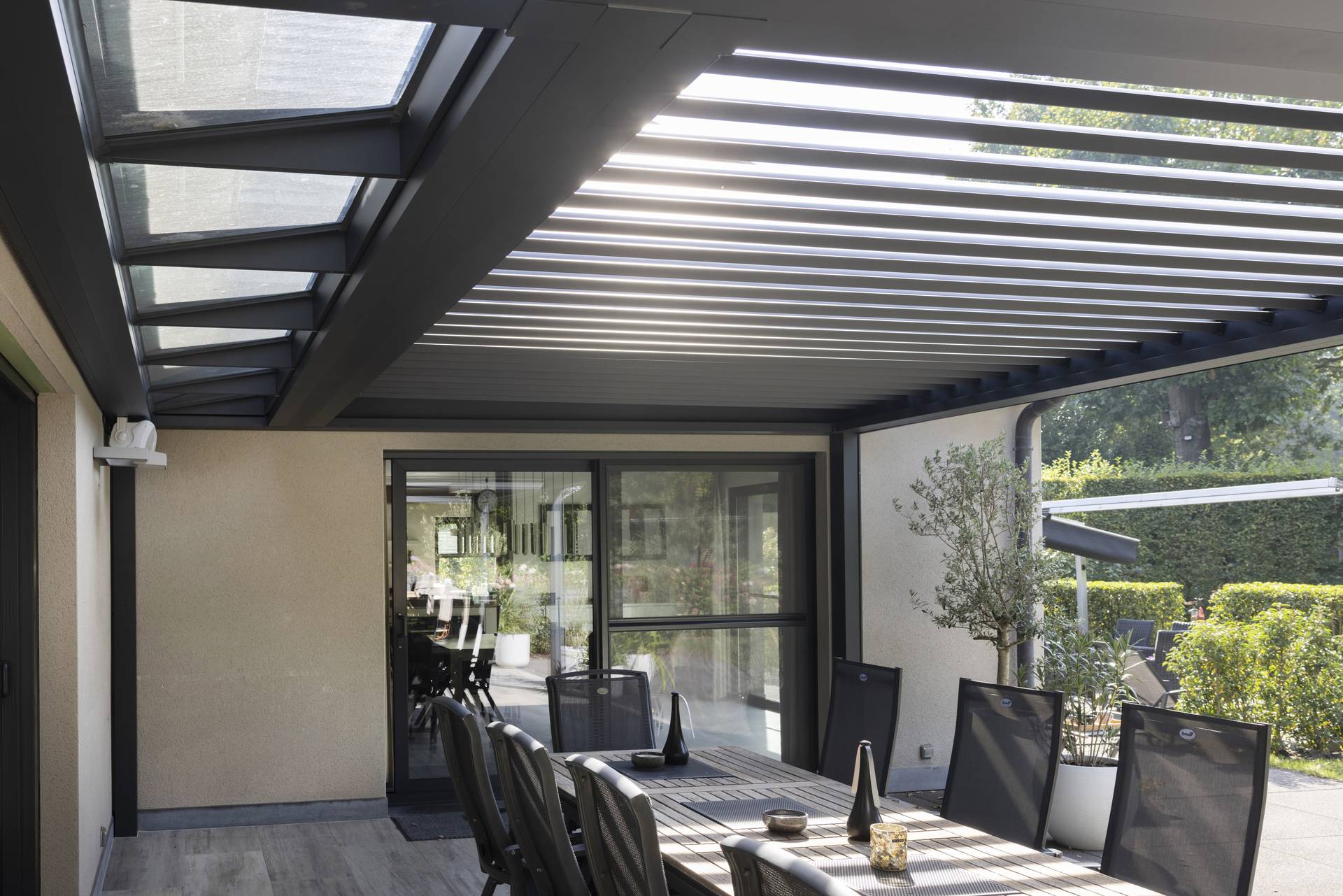Aluminium‑Pergola B720 mit festem Glaselement und teilweise geöffneten XL‑Lamellen über einem modernen Essbereich im Freien in heller Gartenumgebung