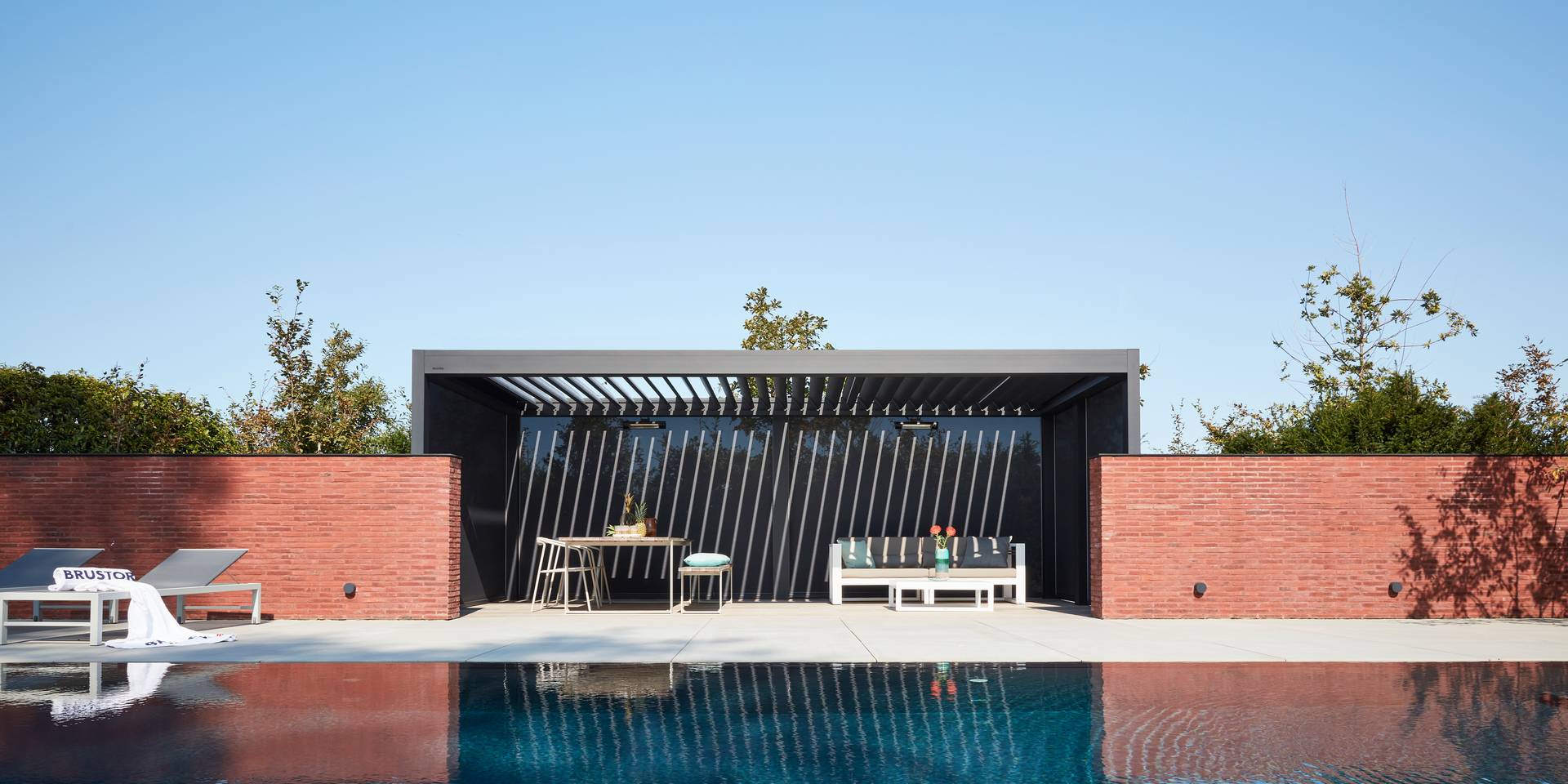 Poolblick auf eine Brustor B200(XL) Lamellenpergola mit verstellbaren Aluminiumlamellen, die eine schattige Outdoor‑Lounge zwischen Ziegelmauern bildet.