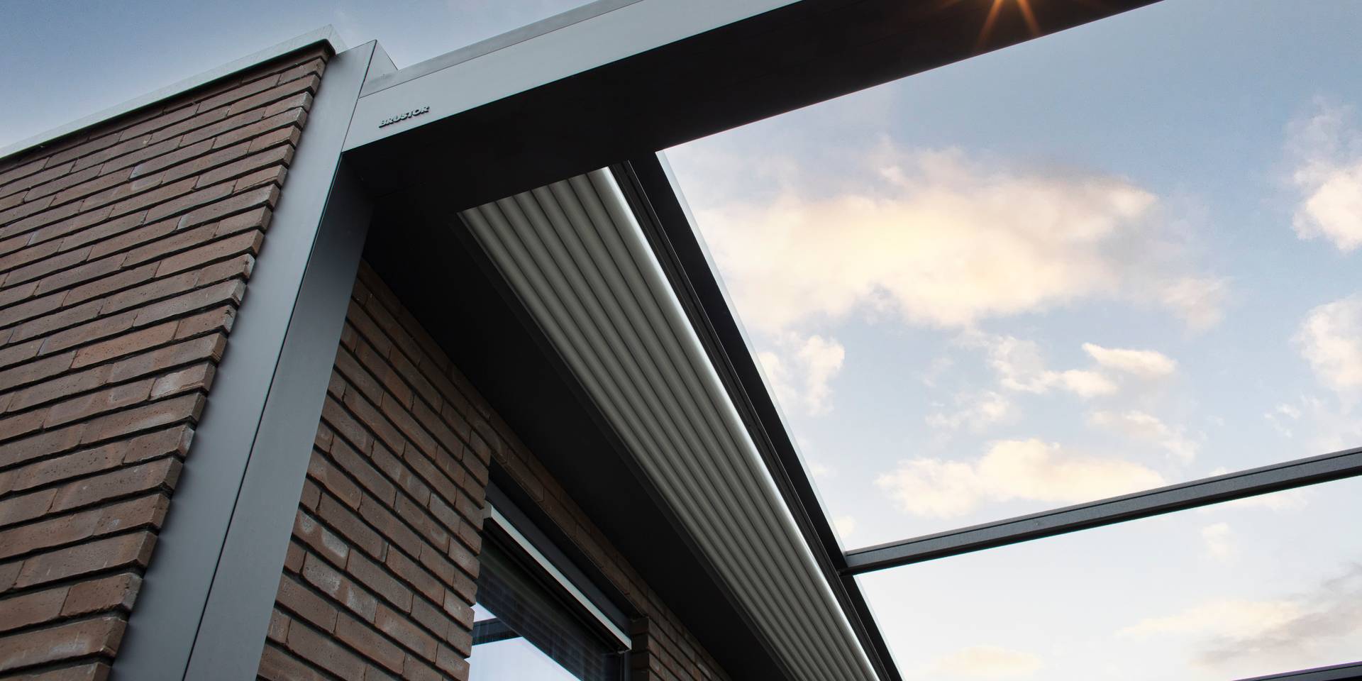 Gros plan d’une pergola en aluminium avec toit en toile rétracté, fixée à une façade en briques, montrant la structure de toiture ouverte vers le ciel.